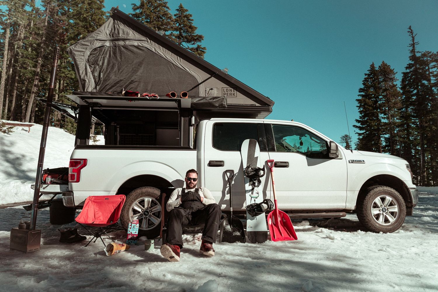 Winter camping setup with white pickup truck, rooftop tent, and ski gear in snowy mountain setting.
