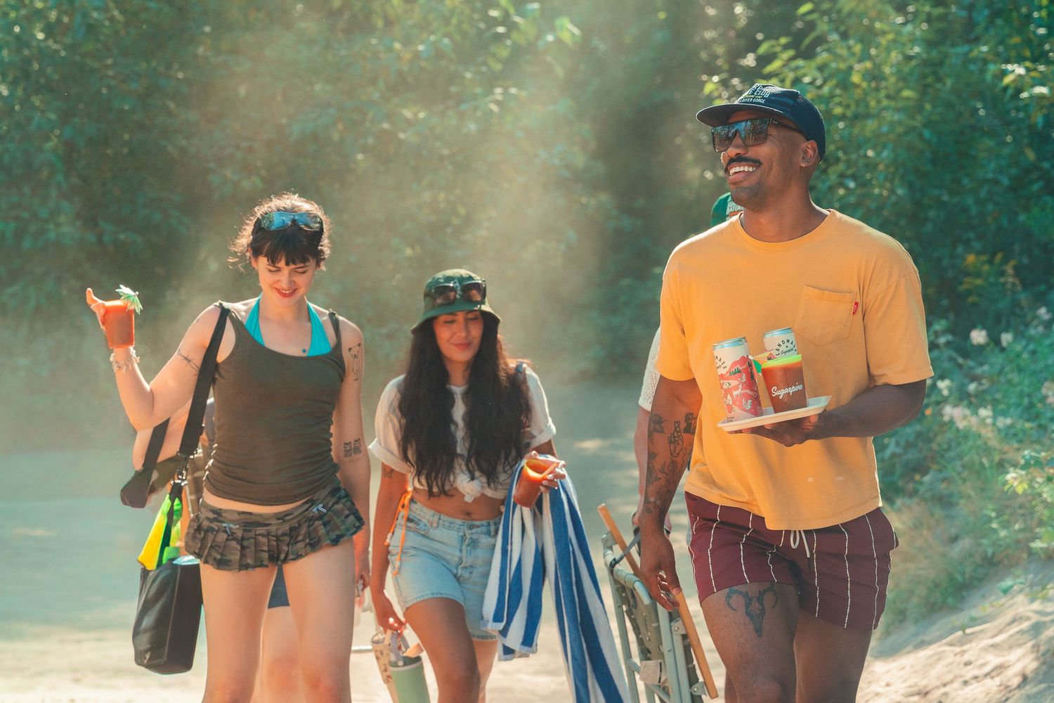 Three friends walk on a sunny dirt path, carrying drinks and bags, smiling in casual summer outfits.