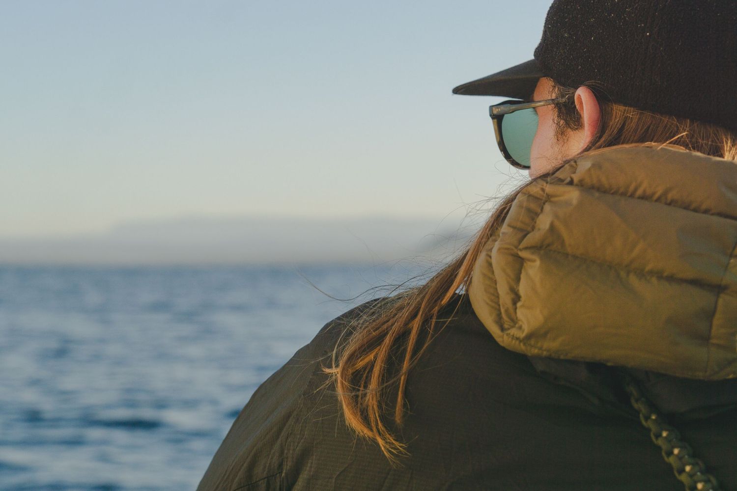 Person in olive puffer jacket and black cap gazes at calm blue ocean horizon on a clear day.