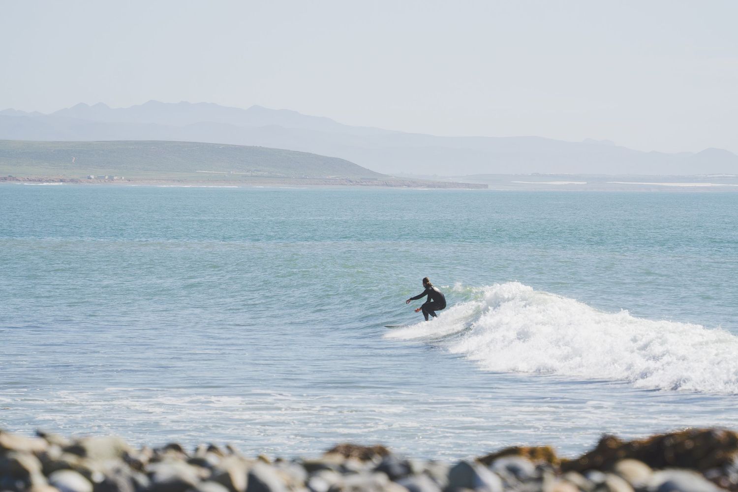 Surfer riding a wave at a rocky beach with mountains visible in the background.