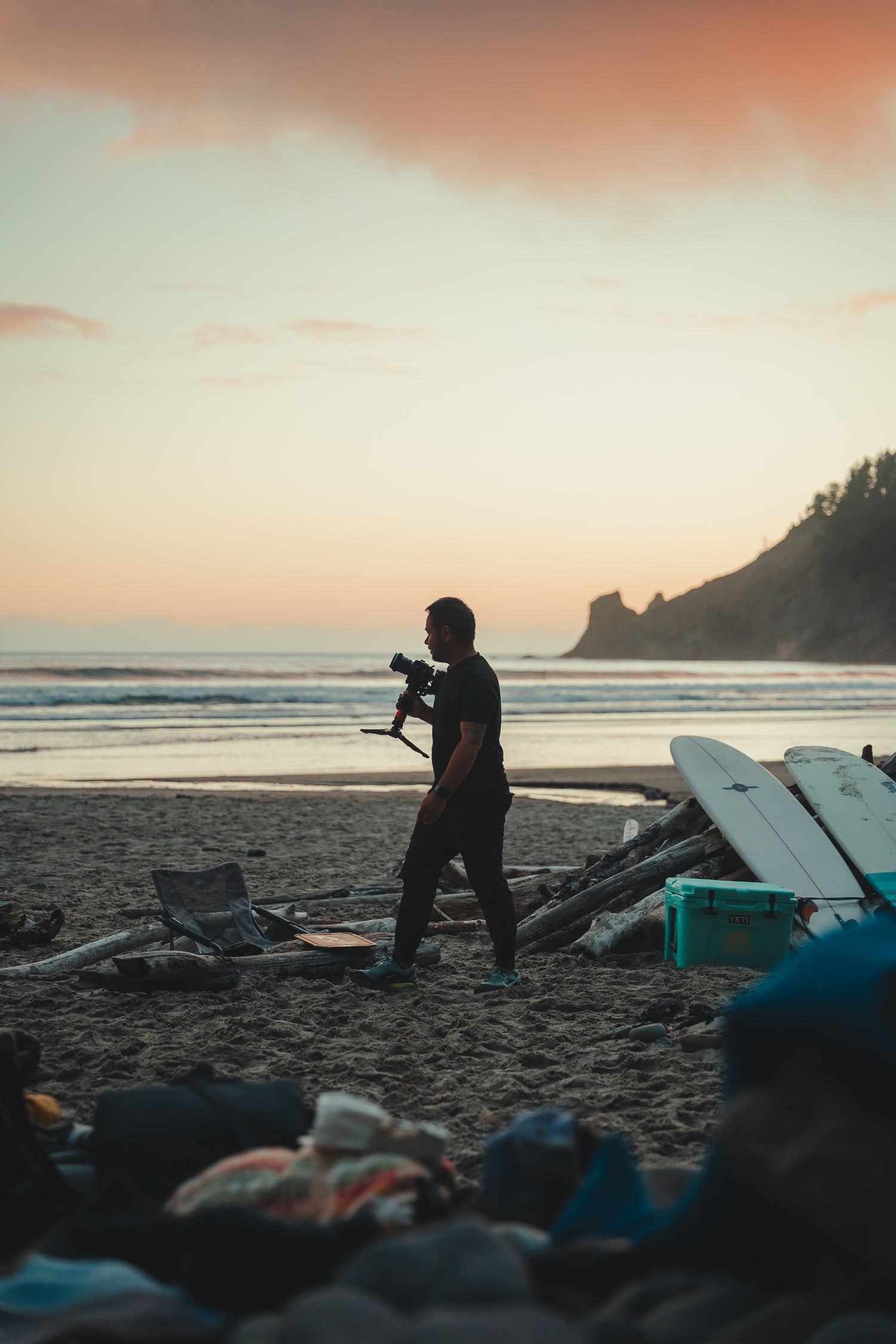 Silhouette of a photographer walking on a sandy beach at sunset with surfboards and a rocky headland in the background.