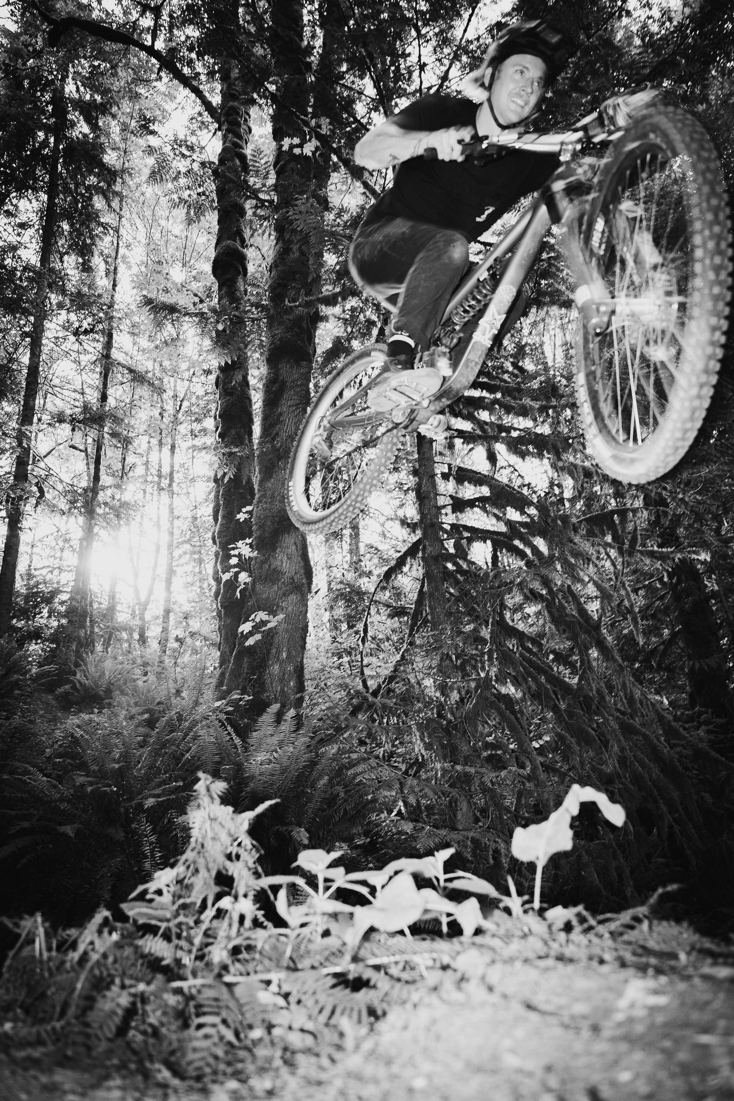 Black and white photo of a mountain biker performing an aerial trick in a forest trail surrounded by trees.