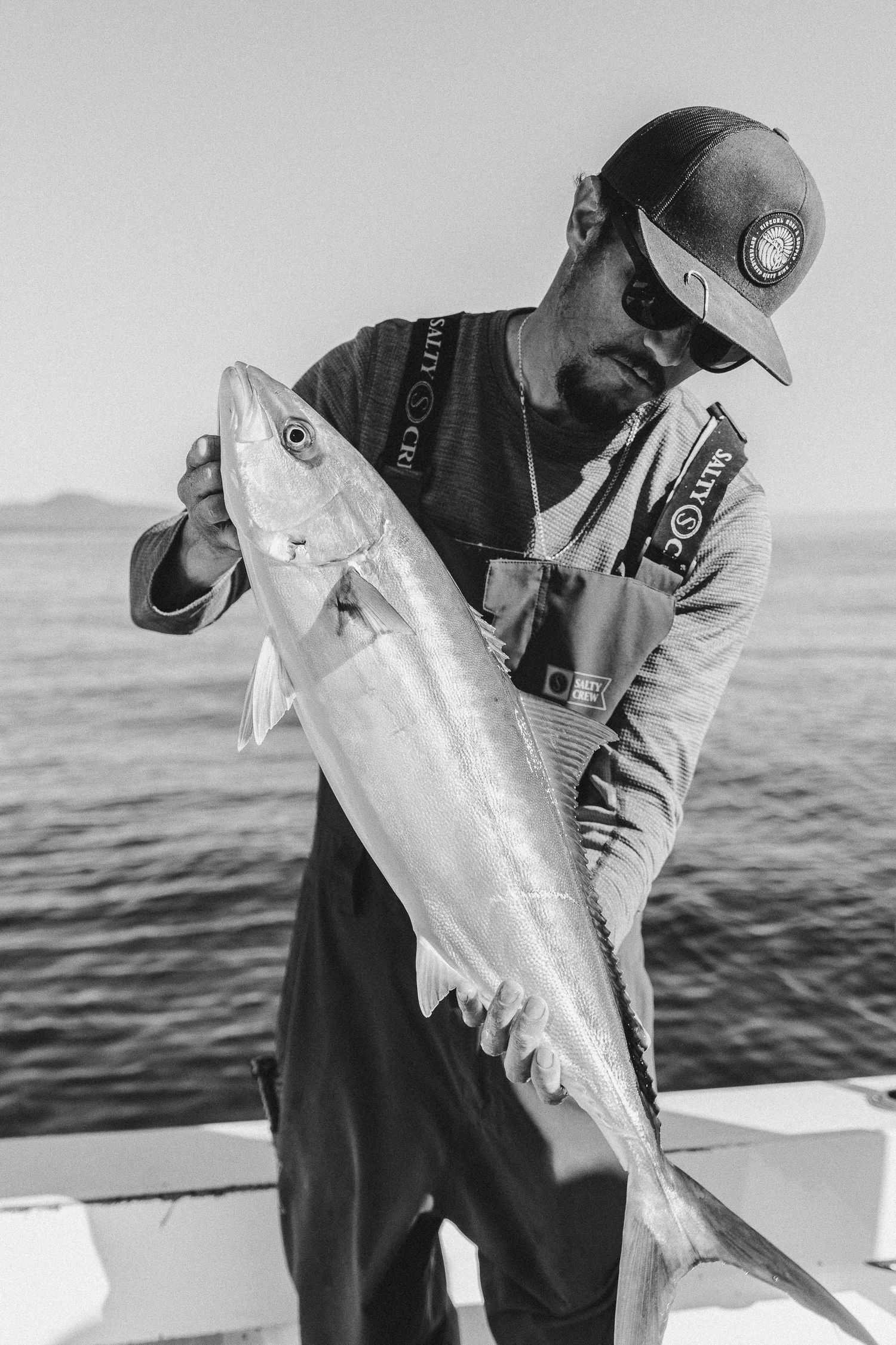 Black and white photo of a fisherman on a boat holding a large caught fish over the ocean.
