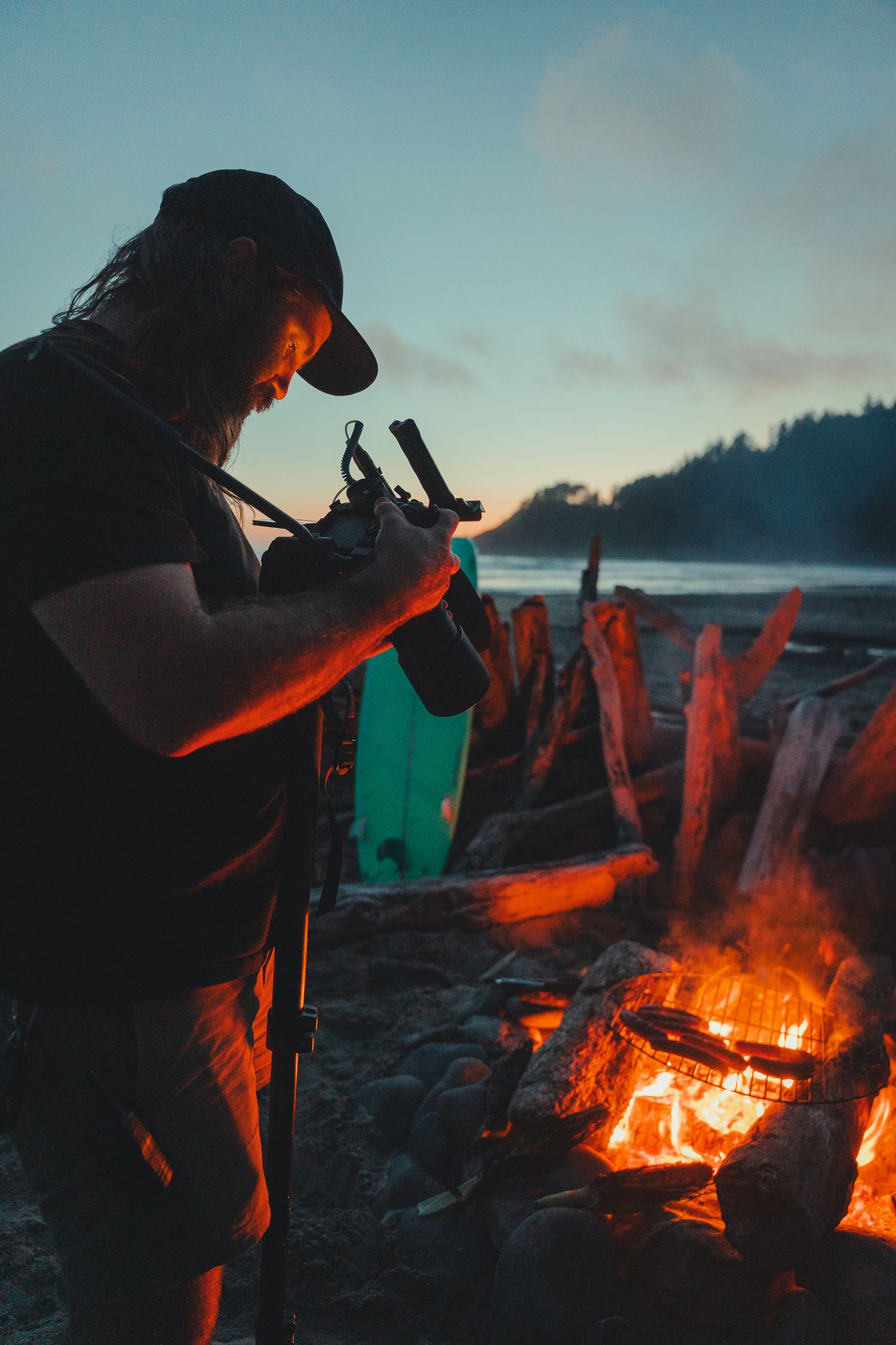 Man holding camera gear beside a glowing campfire on a rocky beach at dusk with misty forest background.