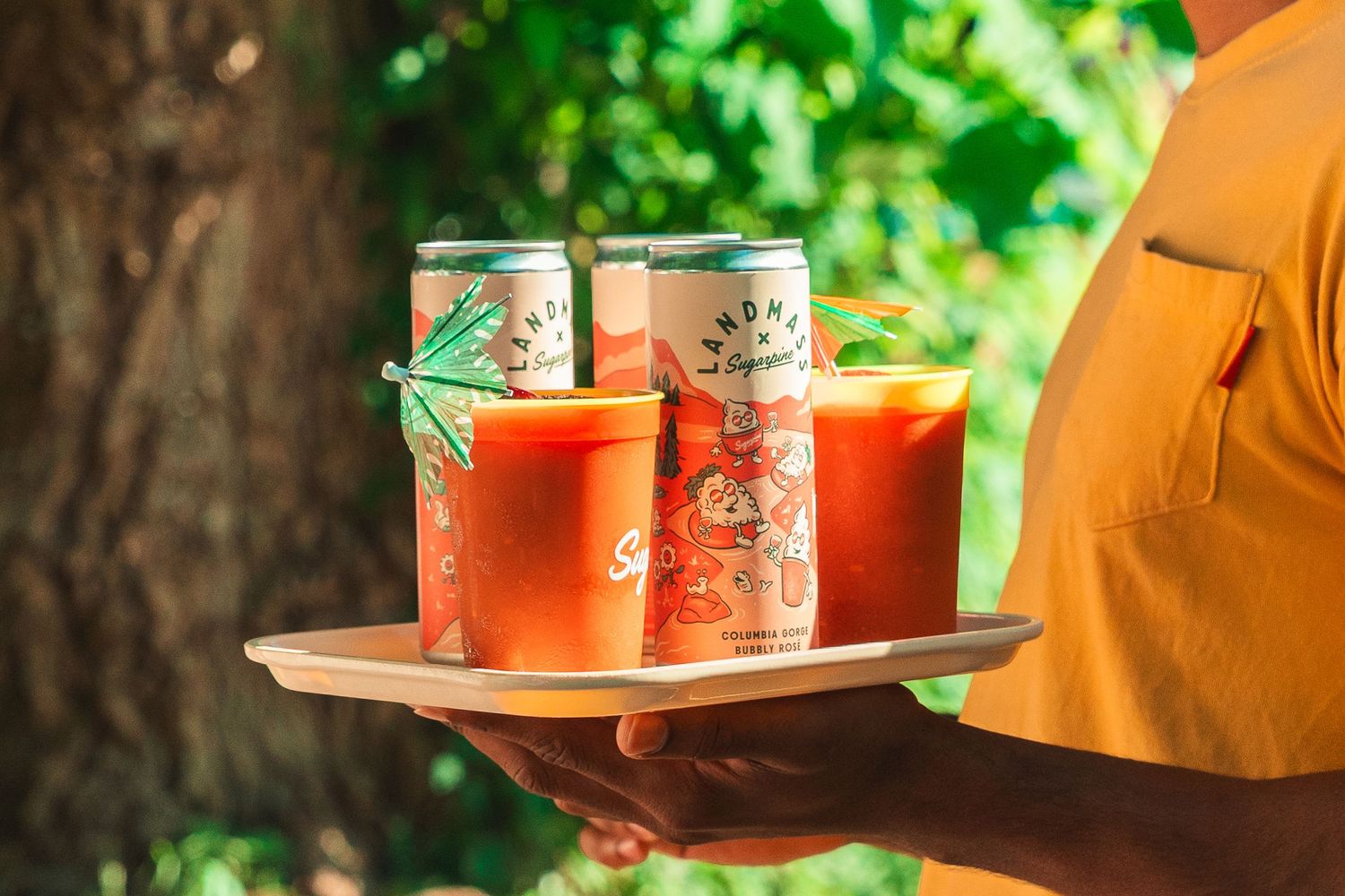 Person holding a tray with Bloody Mary cocktails and canned drinks garnished with green straws outdoors.