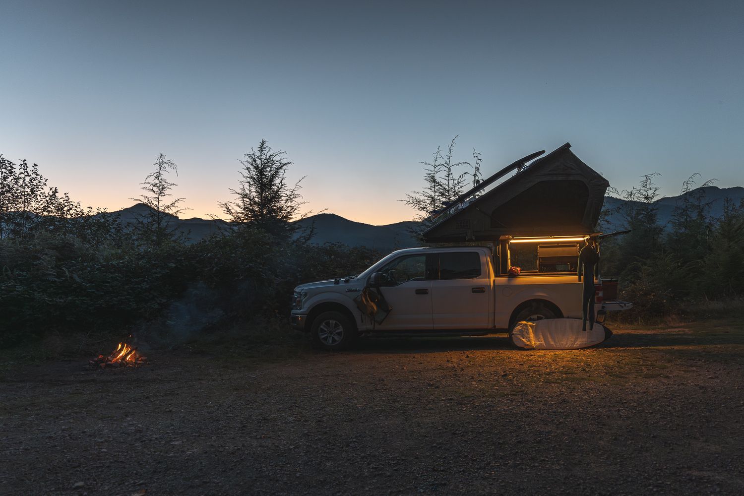 Illuminated camping truck parked in a remote mountain location at dusk with a campfire glowing nearby.