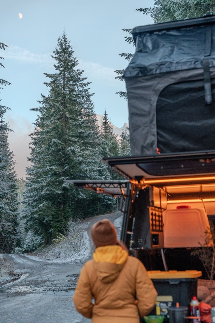 A cozy winter camping setup with snow-covered evergreens and a warm glow from inside a rooftop tent.