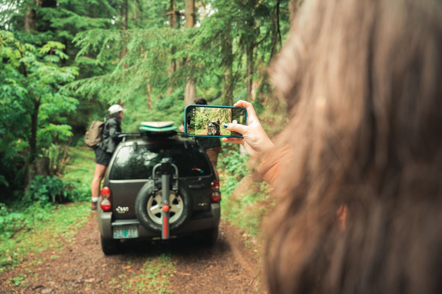 Person photographing a 4x4 SUV with bike rack on a forest trail surrounded by lush green trees.