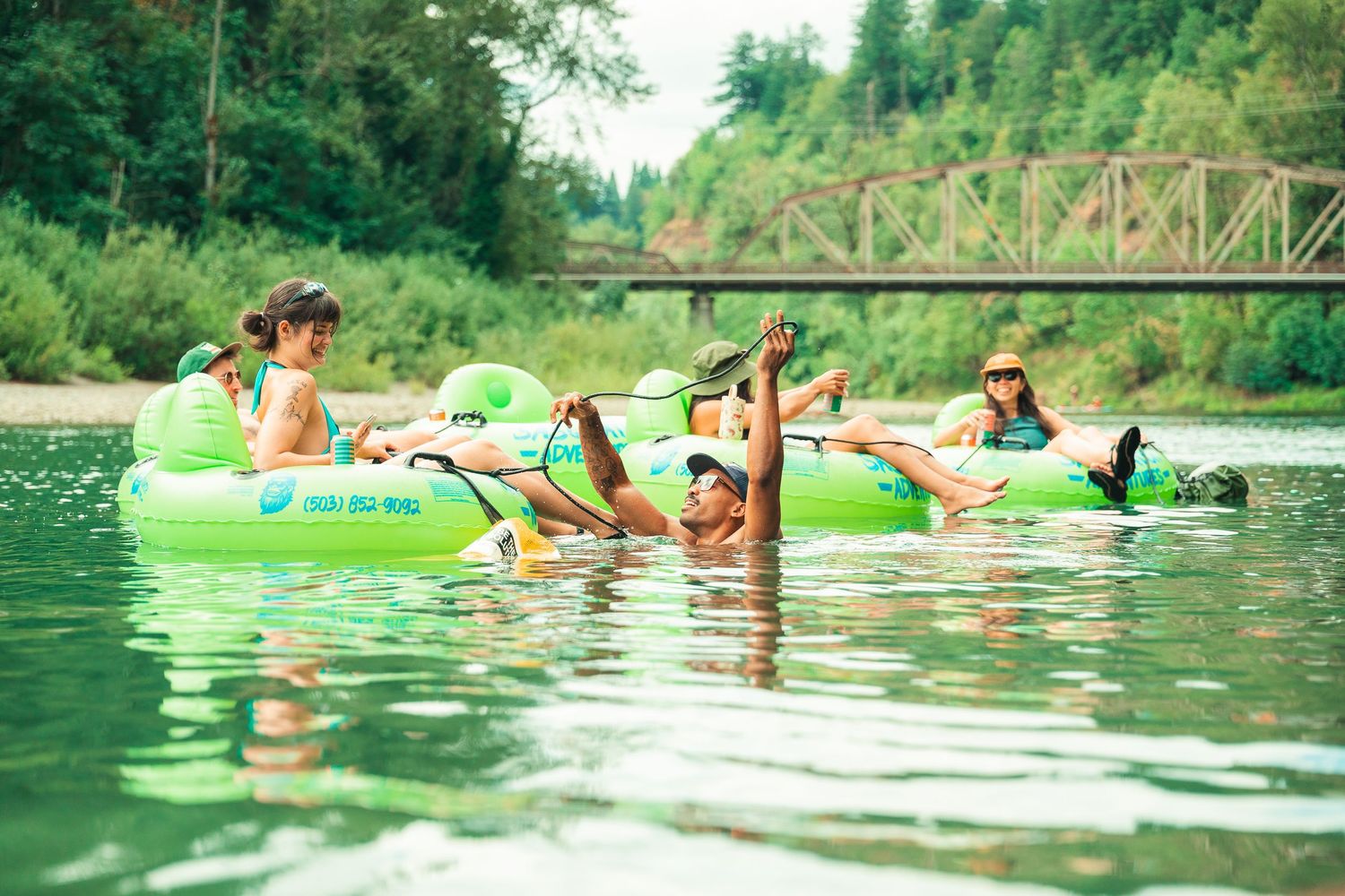 Kids floating on green inflatable tubes in a calm river surrounded by lush trees and a bridge in the background.