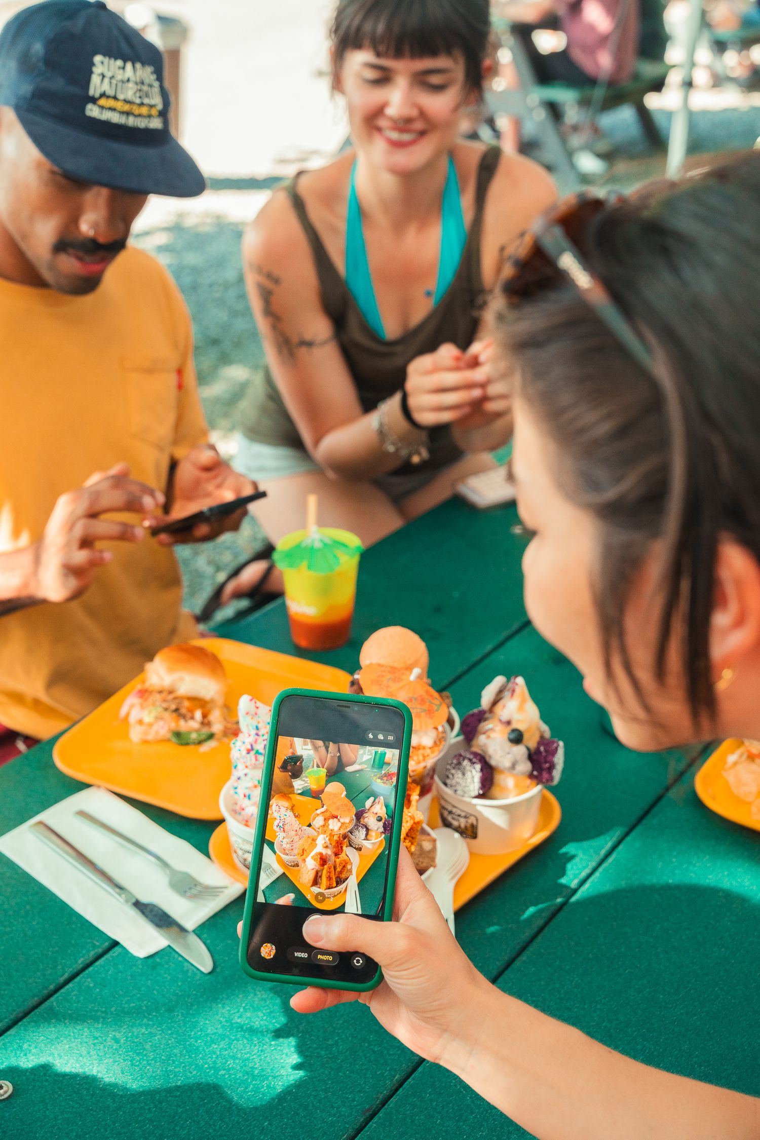 Friends at outdoor table photographing colorful food on smartphone, enjoying sunny day with drinks and sandwiches.