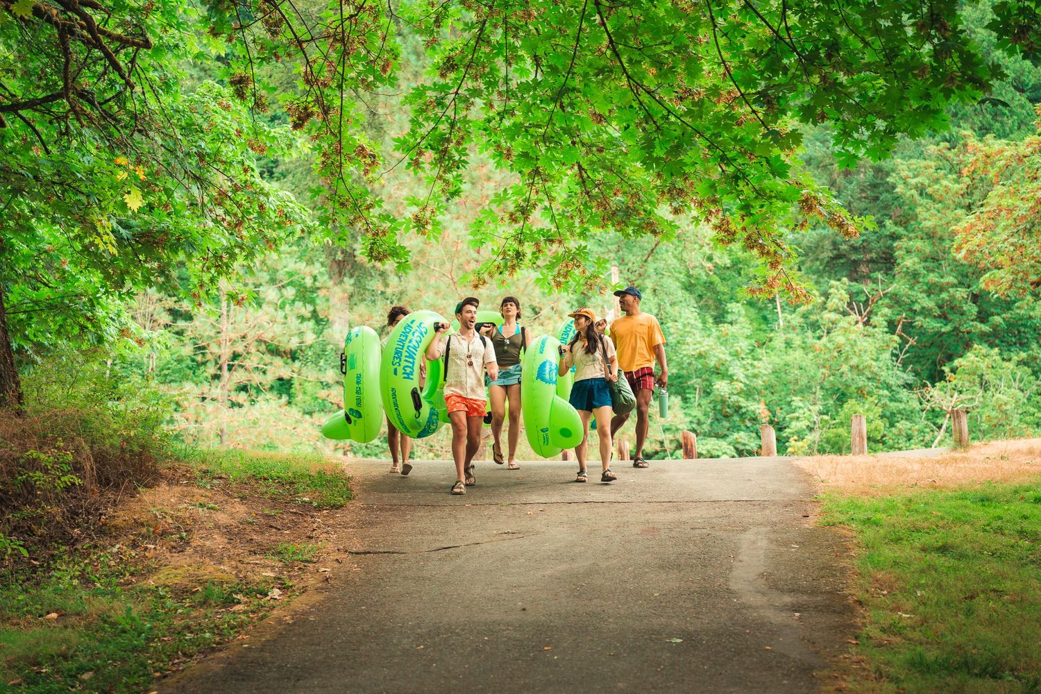 Group of friends walking on a forest path carrying colorful inflatable pool floats on a sunny day.