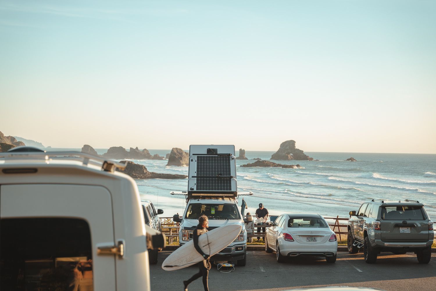 Vehicles parked at a scenic coastal overlook with rocky ocean formations visible in the distance at sunset.