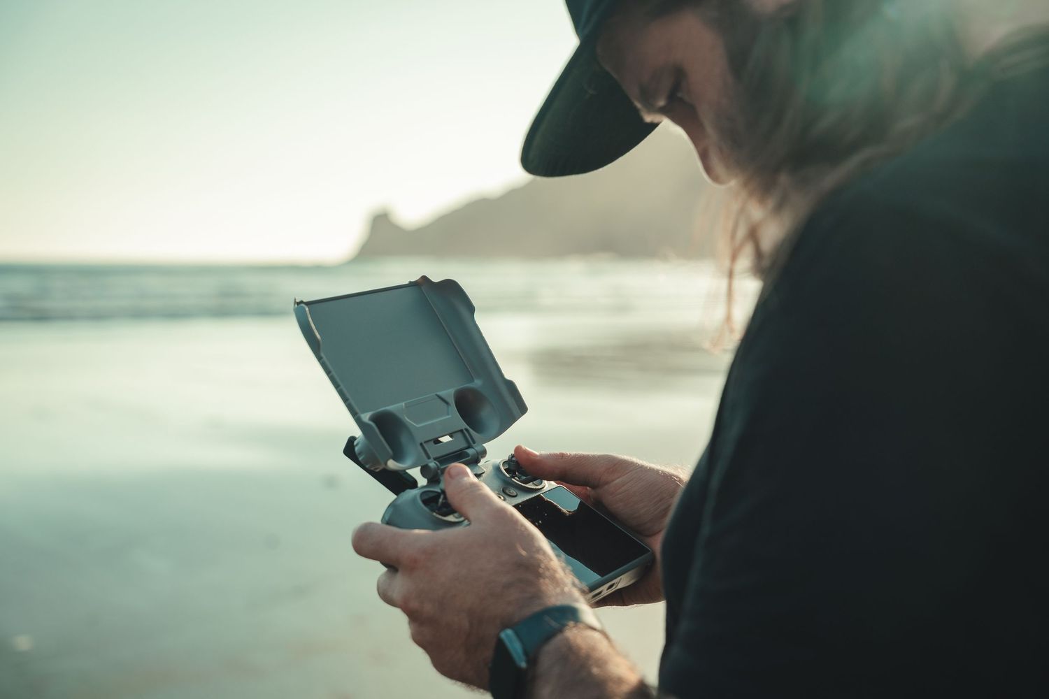 Person in black cap operating a DJI drone remote controller on a scenic beach with ocean backdrop.
