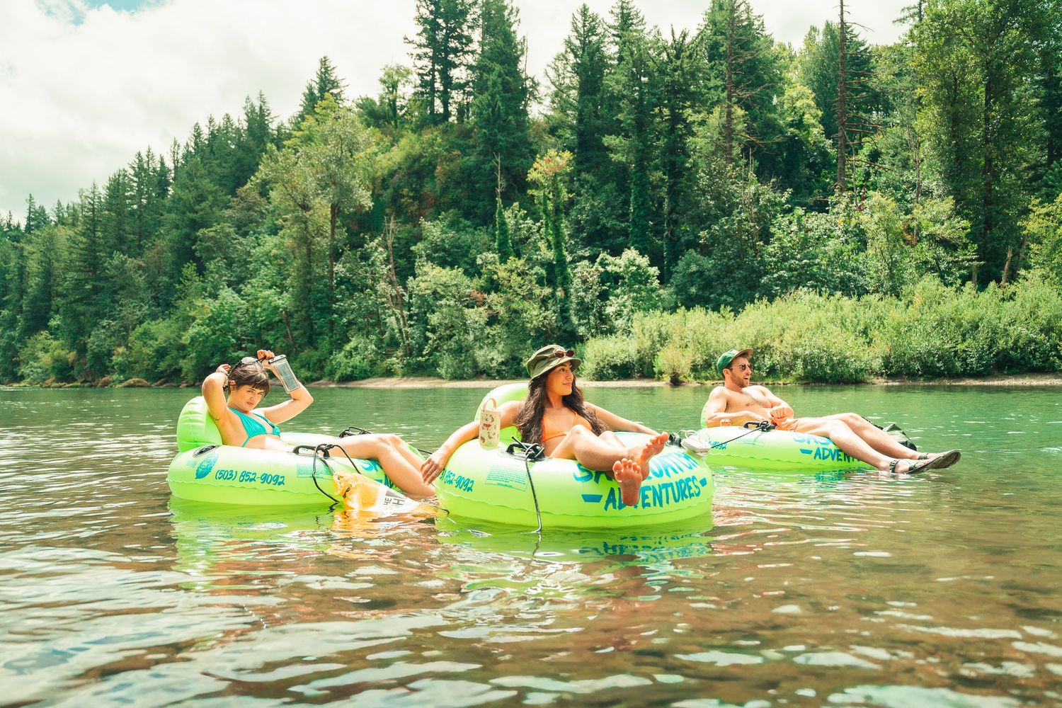 Three people float down a scenic river on green inflatable tubes surrounded by lush forest trees.