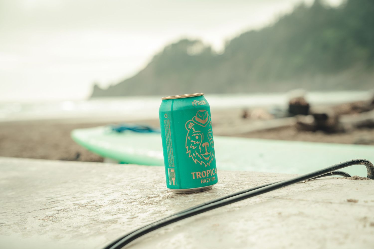 Tropical beer can on a surfboard at a misty beach with green hills in the background.