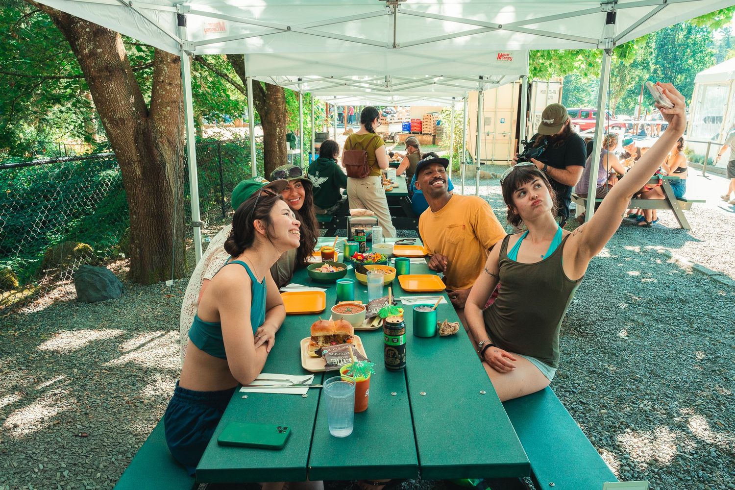 Friends enjoy outdoor dining under a tent, taking a selfie at a green picnic table with food and drinks.