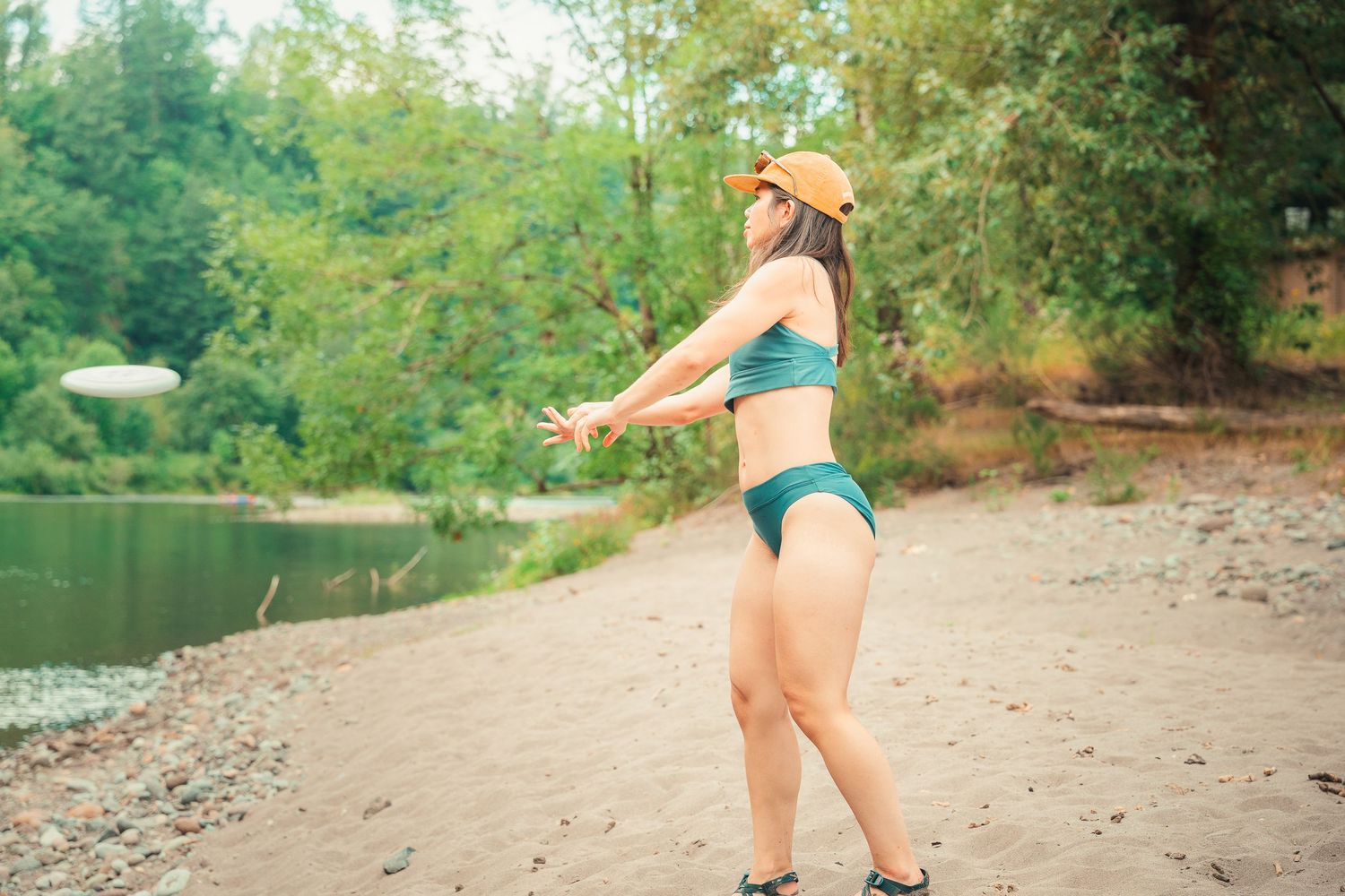 Woman in green bikini and orange cap throwing a frisbee on a sandy beach surrounded by lush trees.