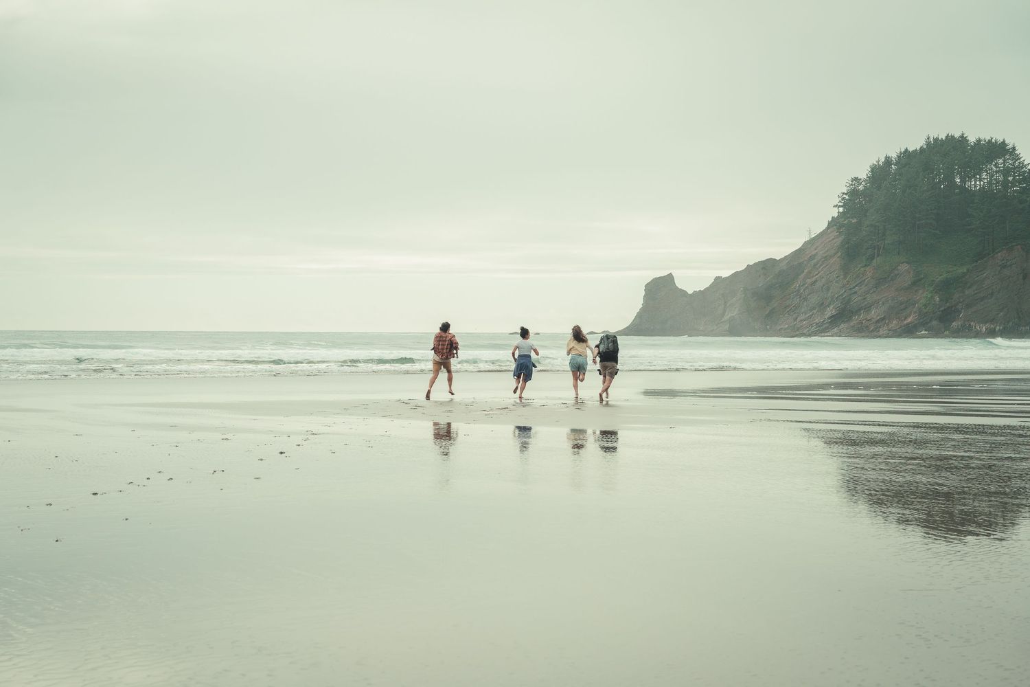 Four people walk along a misty beach shoreline with rocky cliffs and calm ocean waves in the background.