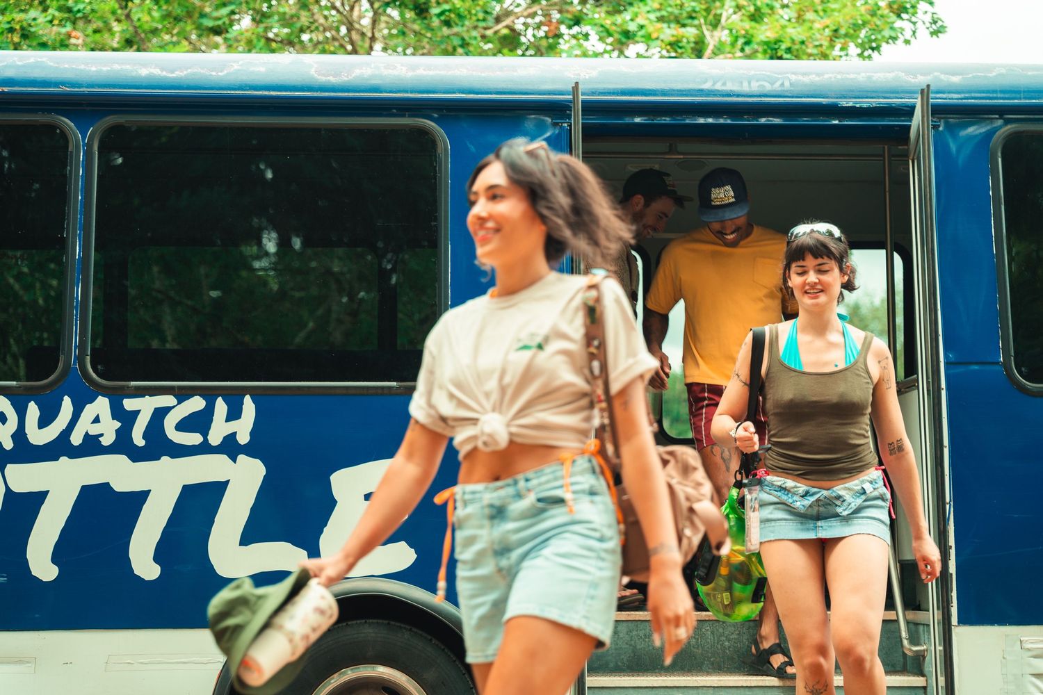 Smiling woman exits a Sasquatch Shuttle bus with other passengers in a lush outdoor setting.
