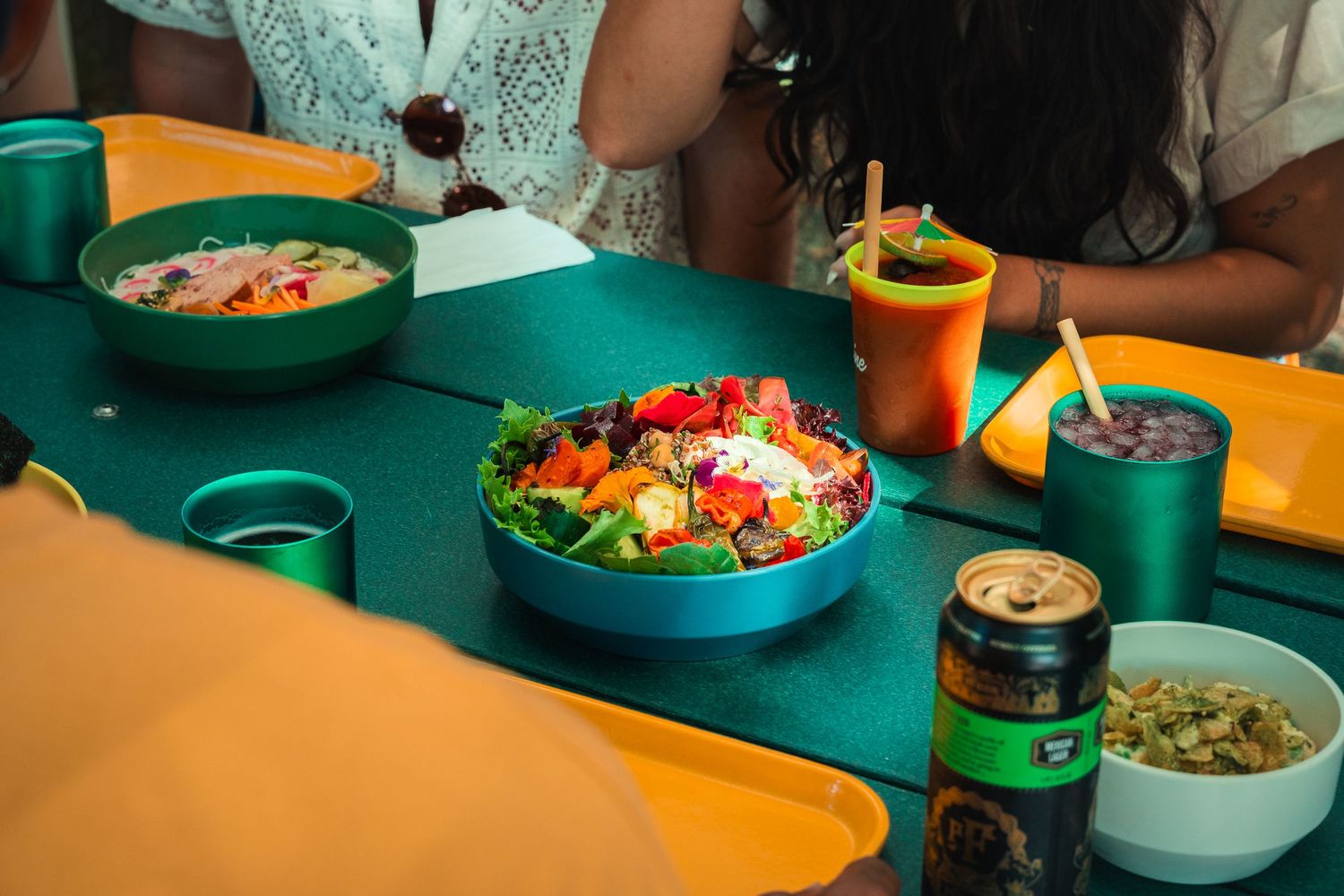 Colorful bowls of fresh salad and drinks on a green tray at a casual dining table with people seated nearby.