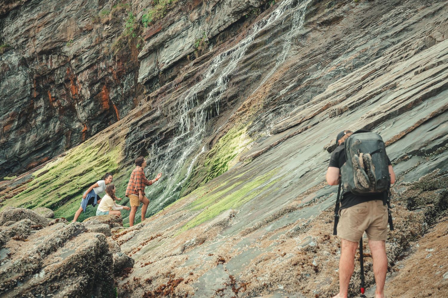 Three hikers with backpacks climbing a steep rocky cliff face with colorful green and orange mineral streaks.