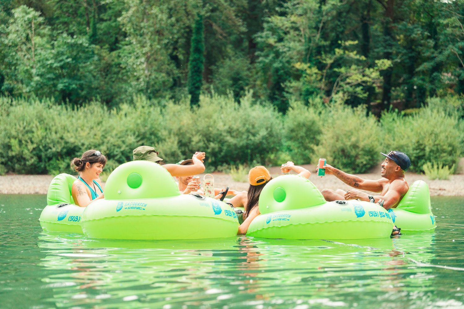 People relaxing on green inflatable river tubes floating on calm water surrounded by lush green trees.