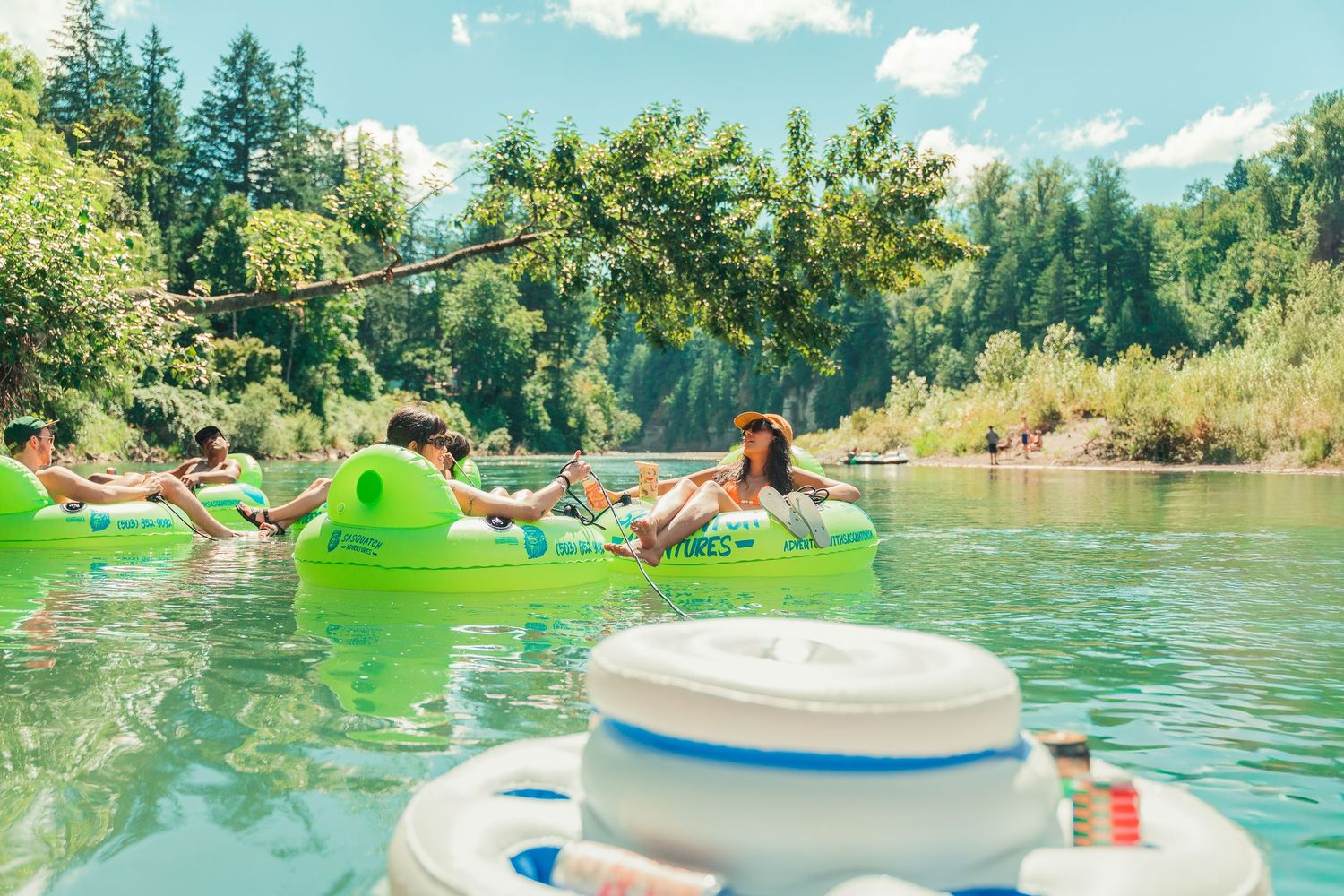 Group of friends relaxing on green inflatable tubes floating down a scenic river surrounded by lush trees.