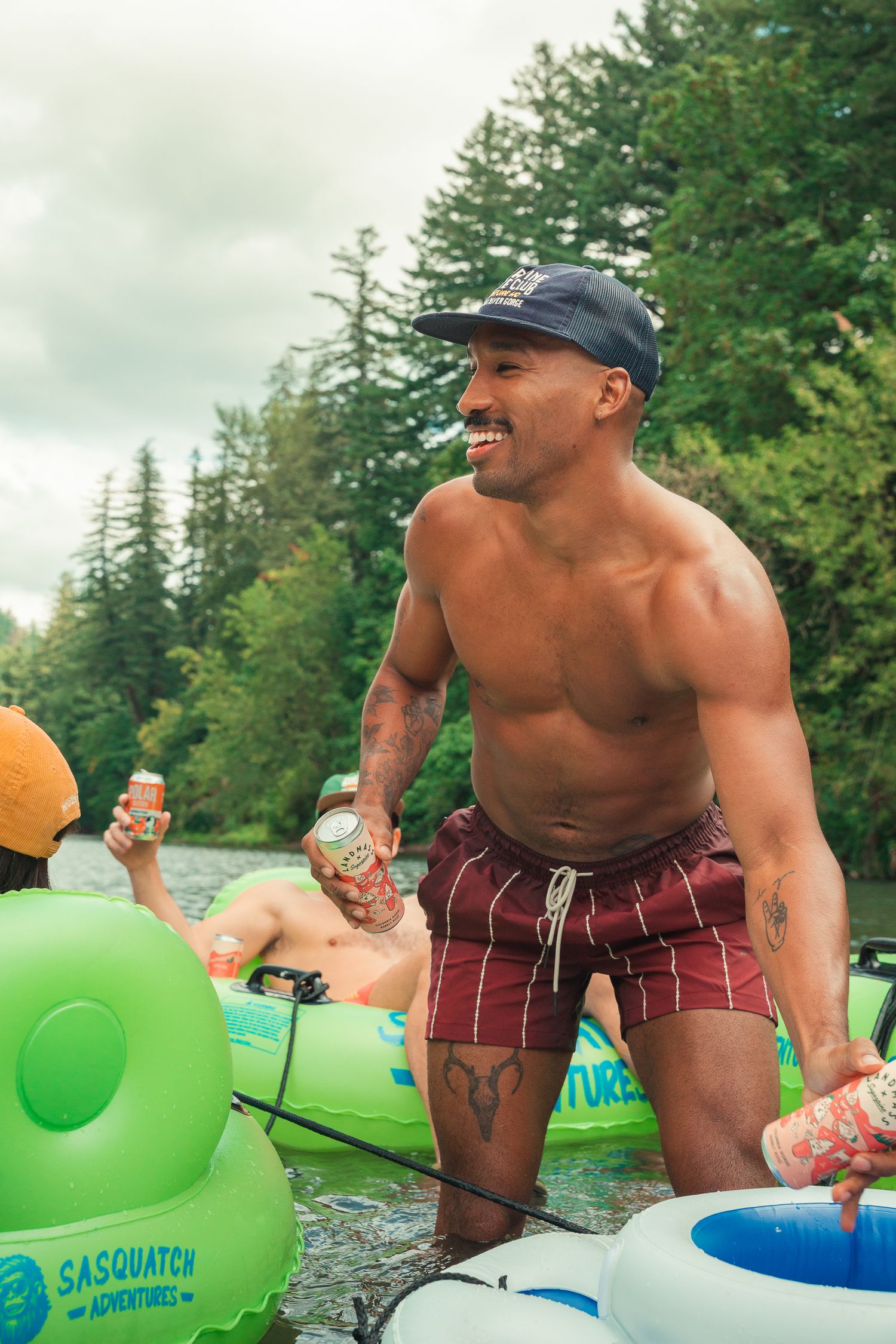 Shirtless man holding drinks on a river float trip with green inflatable tubes and Sasquatch Adventures branding.