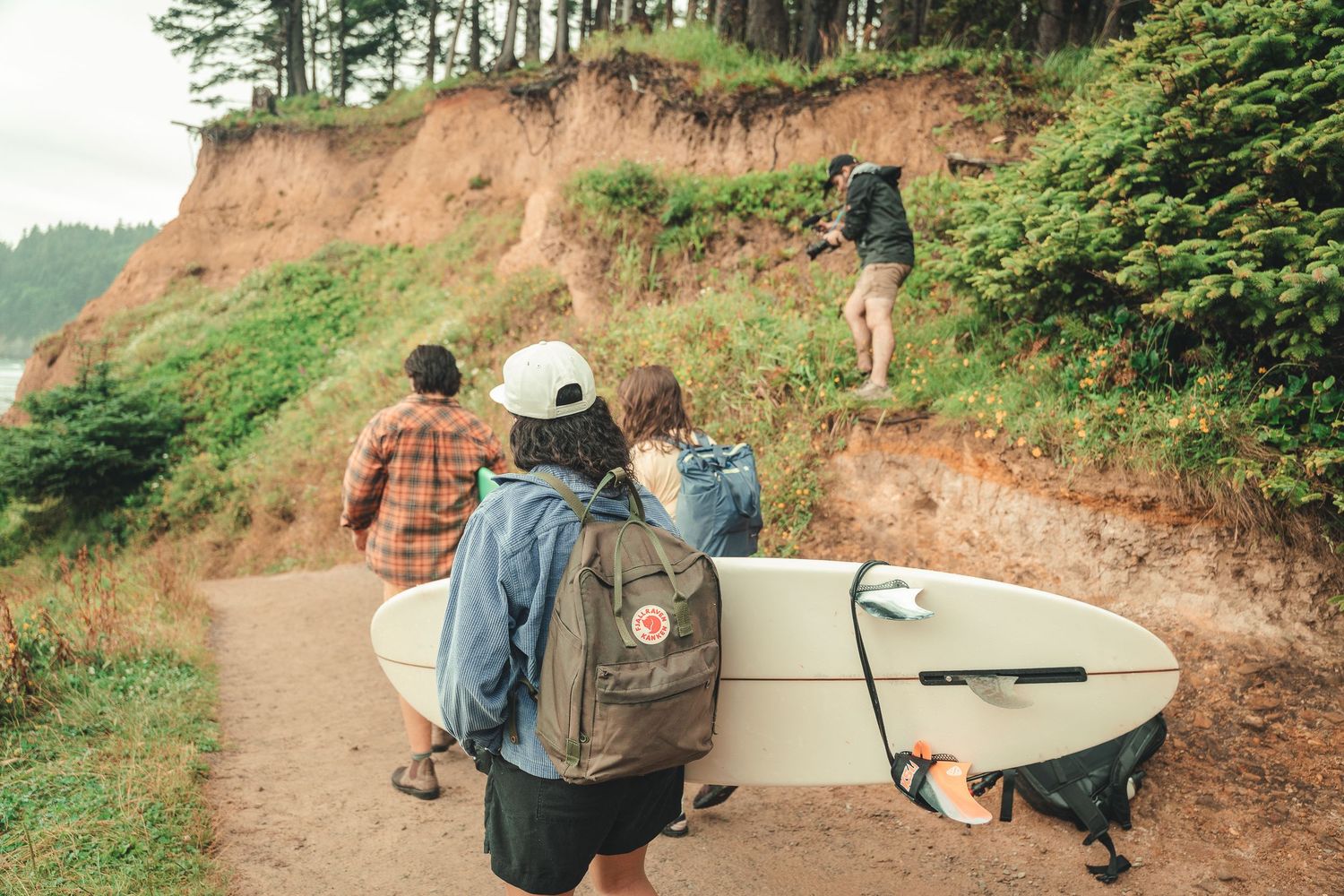 Group of surfers hiking a coastal trail carrying surfboards and backpacks with red clay cliffs and trees in background.