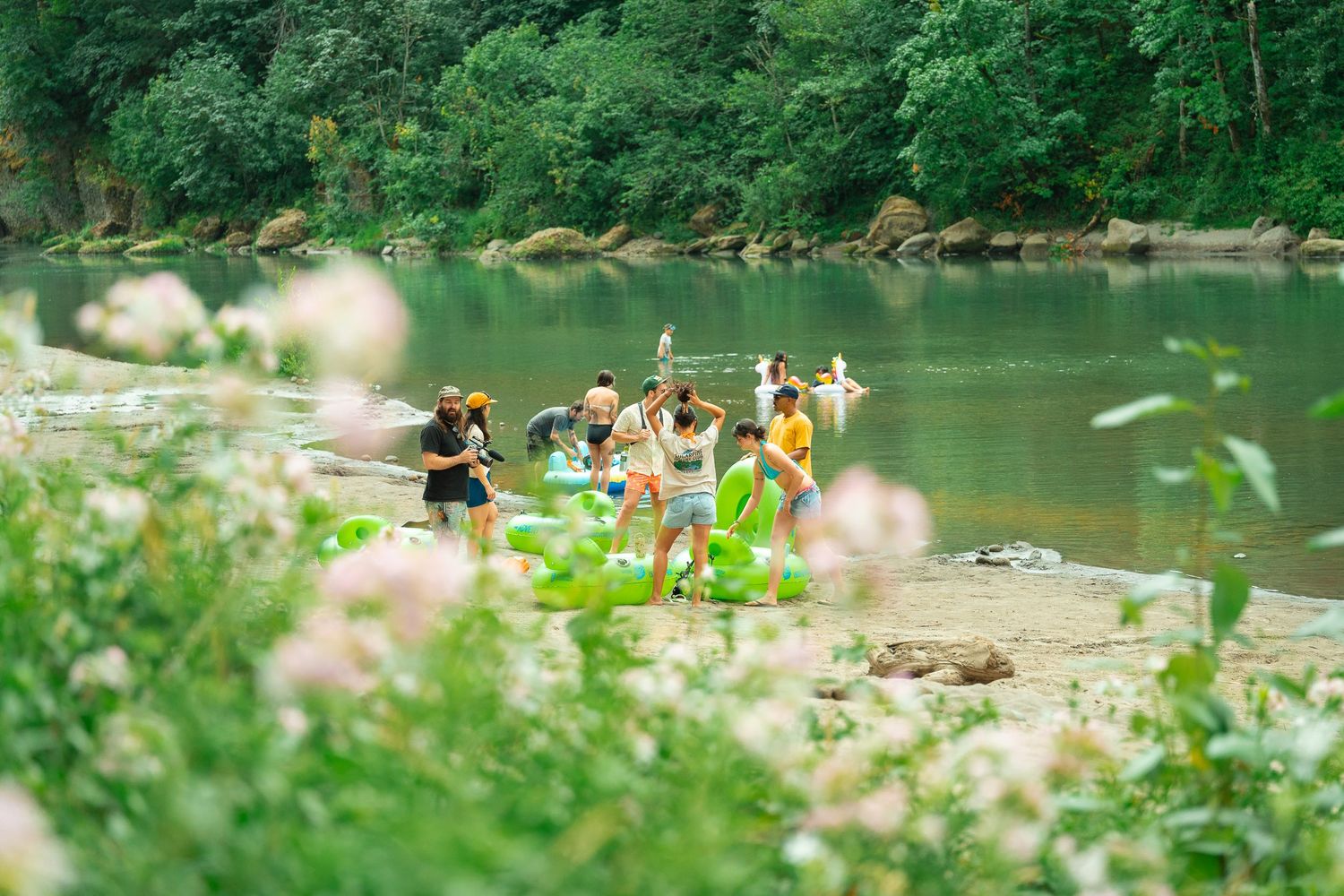 Group of friends enjoying a riverside adventure, surrounded by lush green trees and wildflowers on a sunny day.