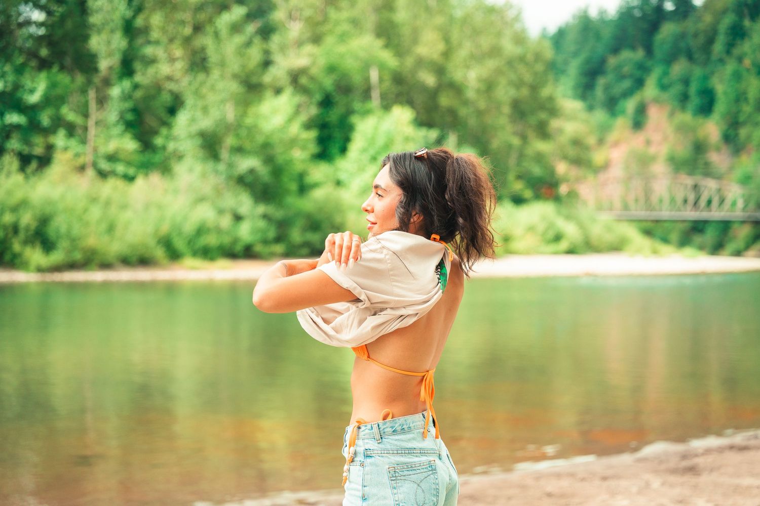 Young woman in crop top and denim shorts stands by a river surrounded by lush green trees on a sunny day.