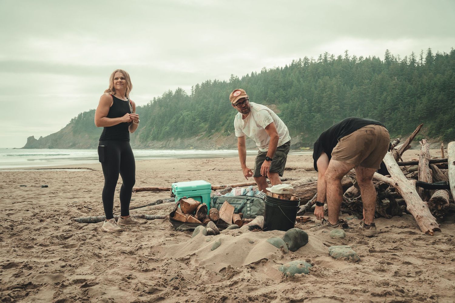 Three people preparing food on a sandy beach with driftwood, ocean waves, and forested hills in the background.