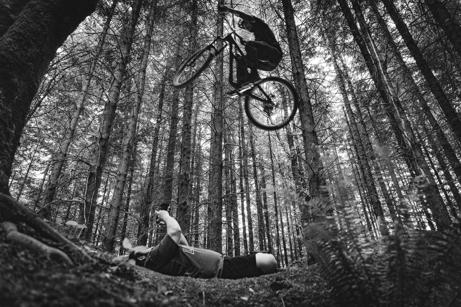 Black and white photo of a mountain biker performing a jump trick above a fallen rider in a dense forest.