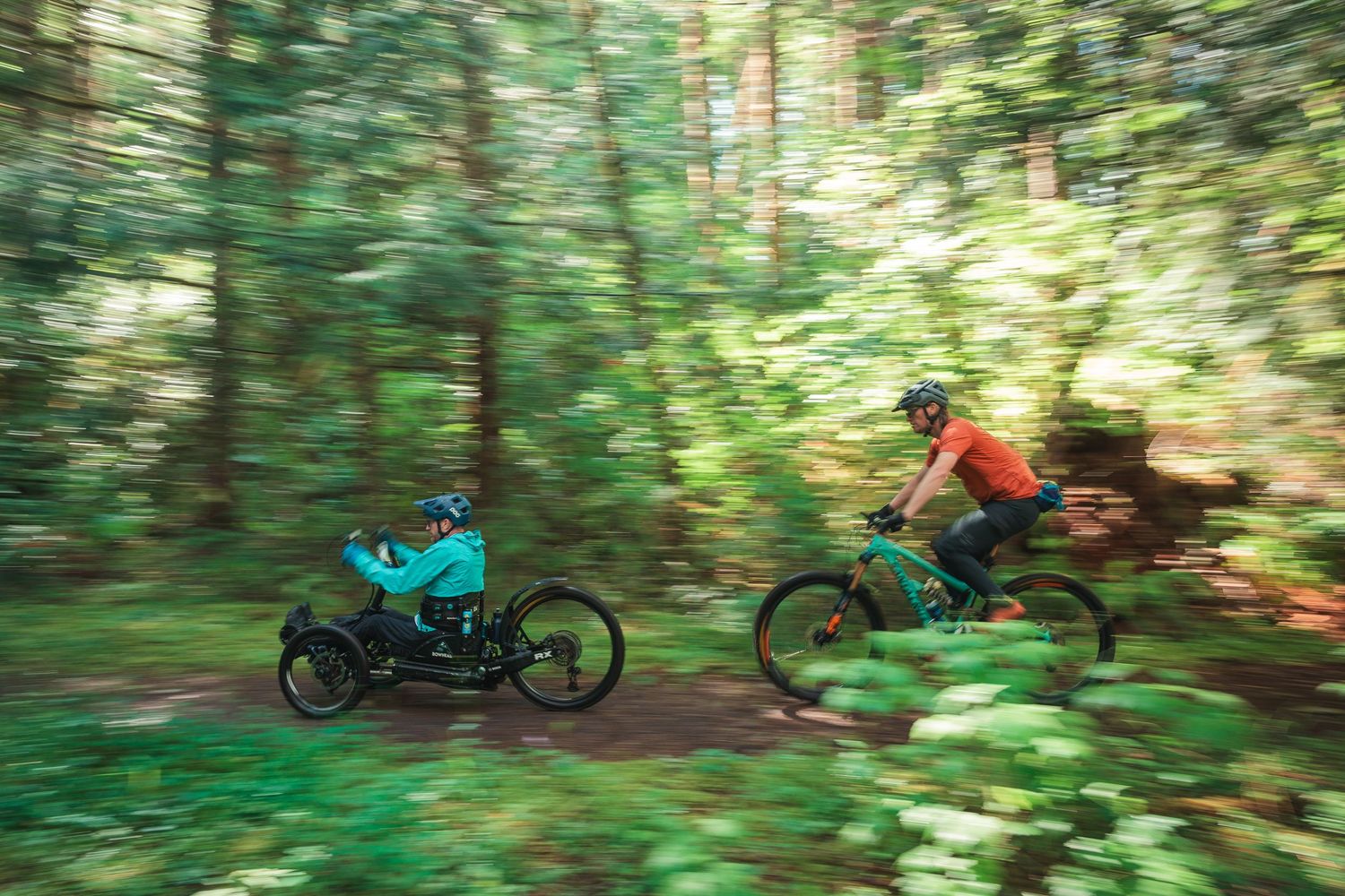 Mountain biker rides through lush green forest trail while towing a child in a blue bike trailer at high speed.