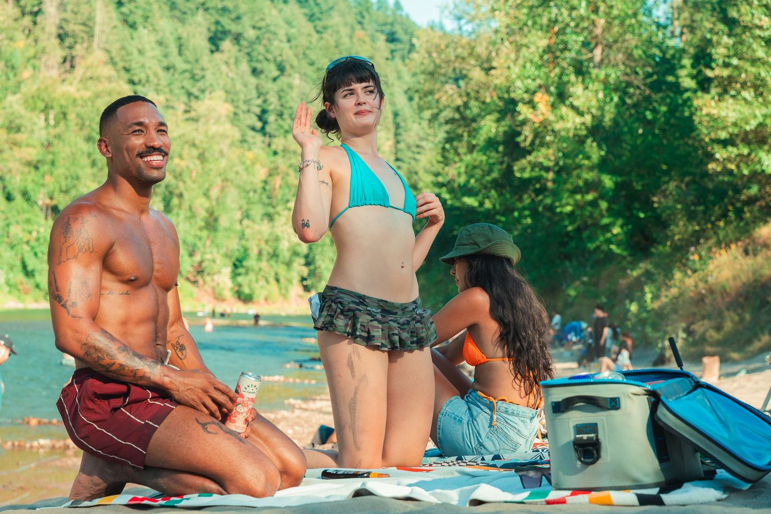 Friends relaxing on a sunny riverbank, with a shirtless man and woman in a teal bikini enjoying a summer day outdoors.