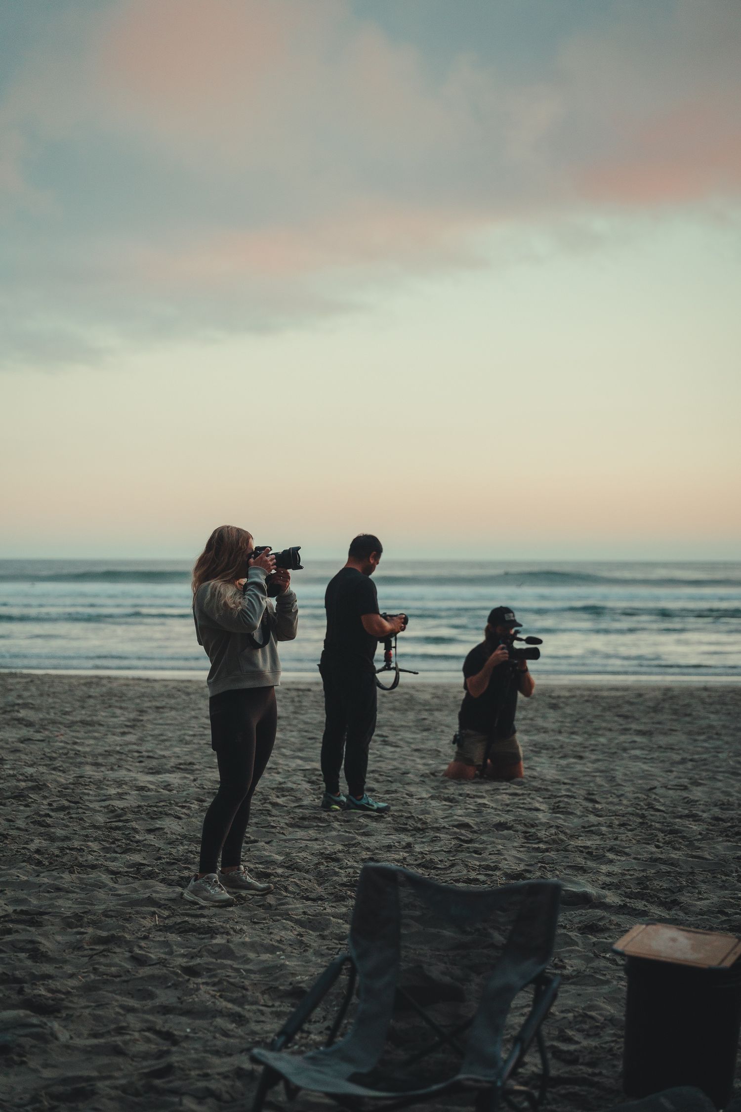 Three photographers capturing ocean waves on a sandy beach at dusk with a pastel sunset sky.