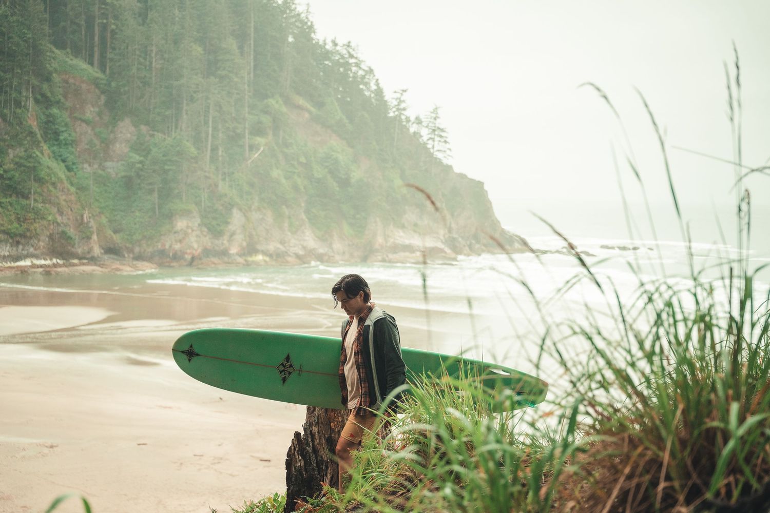 A surfer holds a green surfboard on a misty Pacific Northwest beach surrounded by lush coastal cliffs and tall grass.