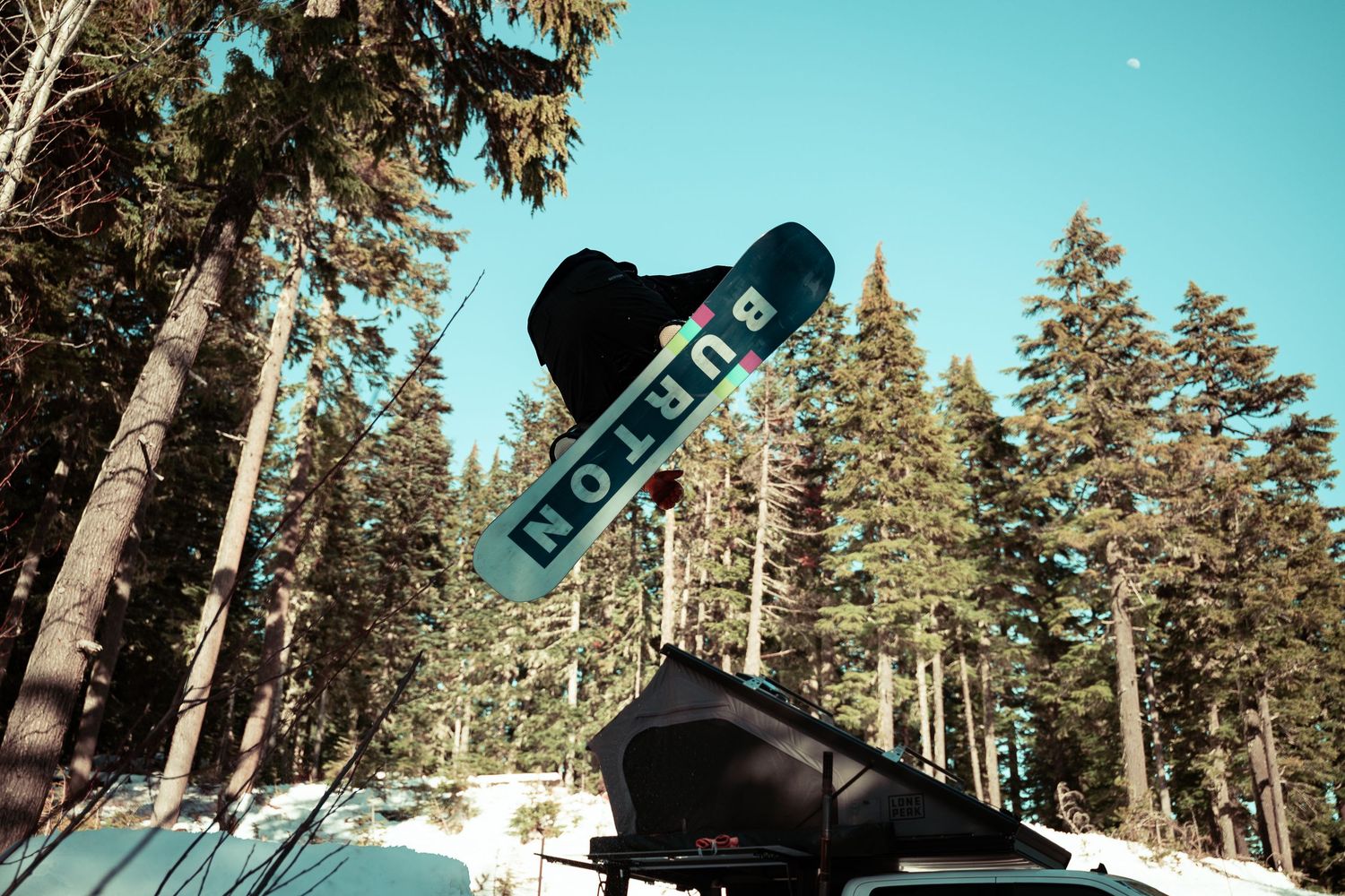 Snowboarder performs aerial jump trick against bright blue winter sky with tall evergreen trees in background.