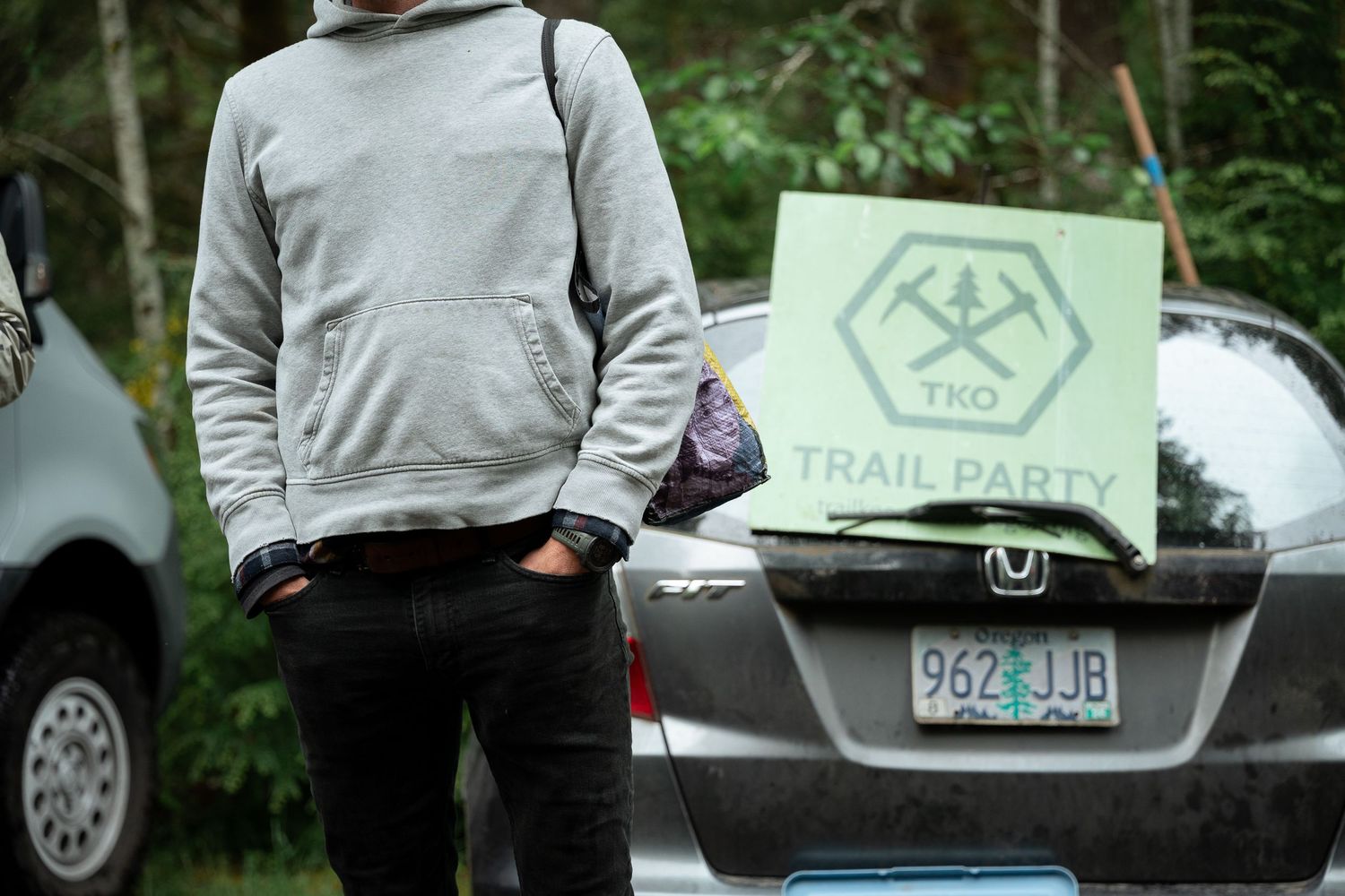 Person in gray hoodie standing by a car trunk displaying a TKO Trail Party sign outdoors.