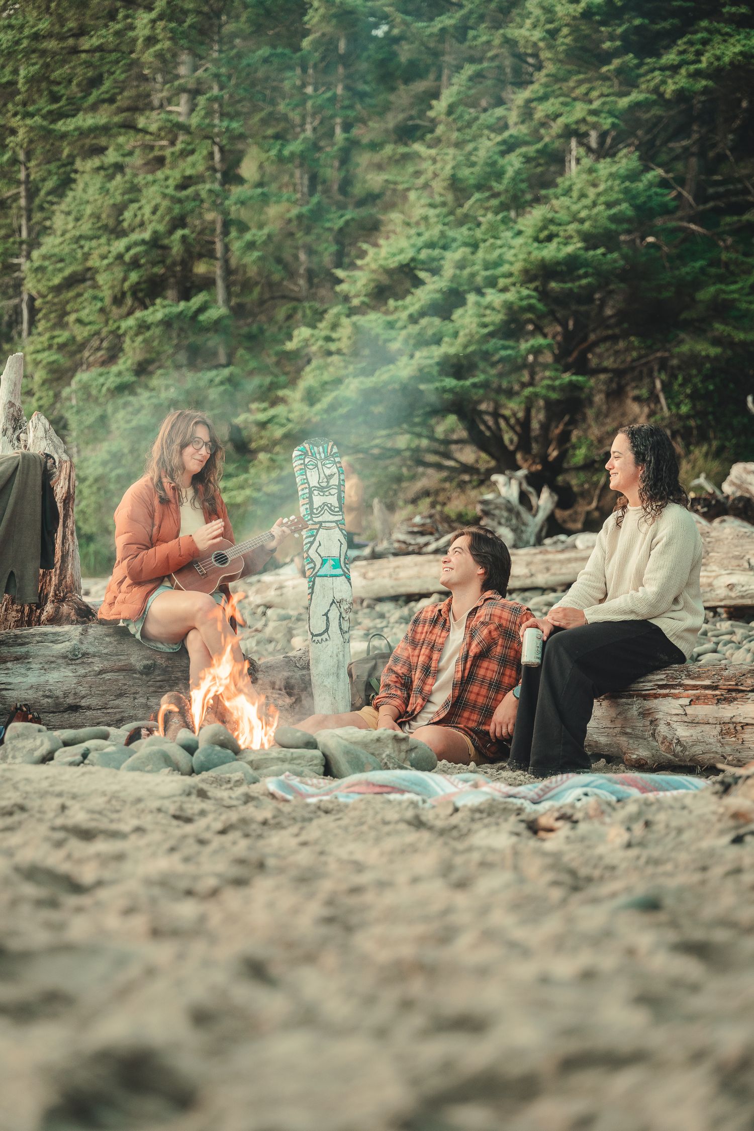 Three friends gather around a campfire on a sandy beach surrounded by lush forest, enjoying a cozy outdoor gathering.