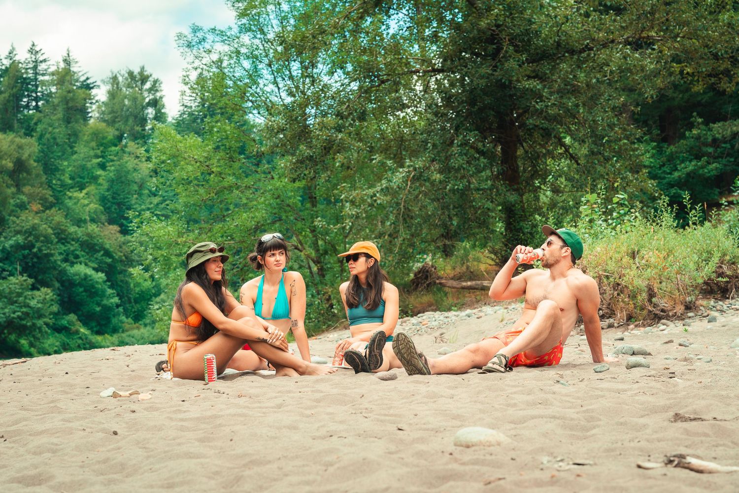 Four friends in swimwear relaxing on a sandy riverbank surrounded by lush green trees on a sunny day.