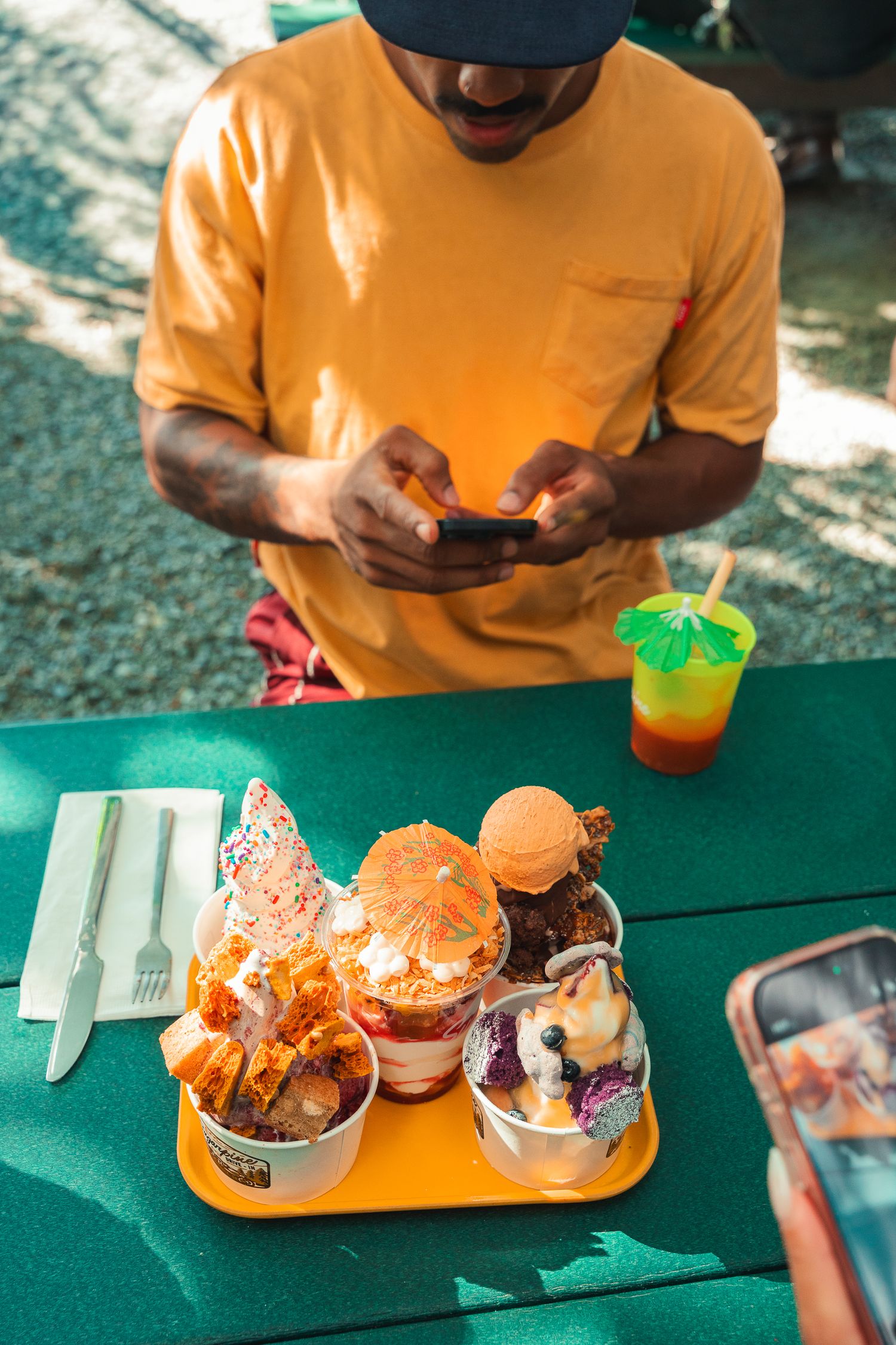 Man in yellow shirt photographing colorful ice cream desserts on orange tray at green outdoor table.