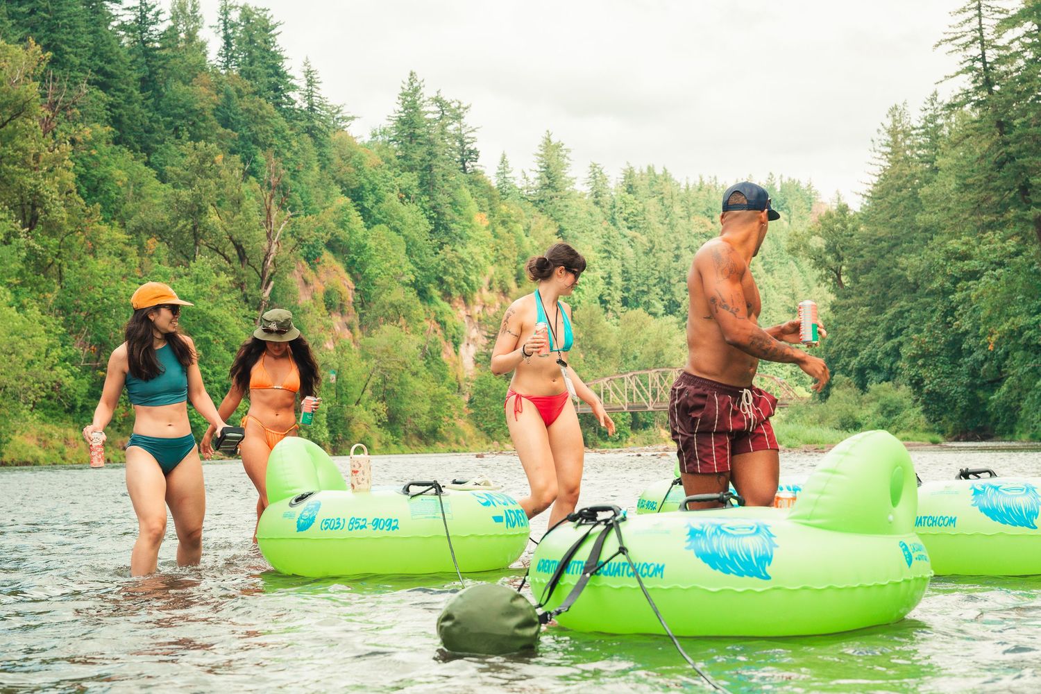 Four friends wade in a river beside green inflatable tubes surrounded by lush forest trees.