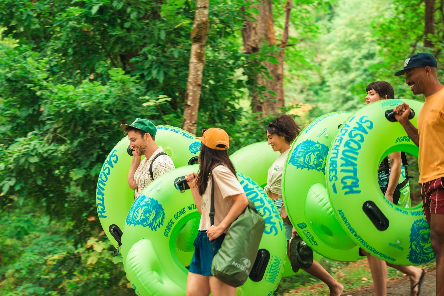 Group of friends carrying green Sasquatch inflatable tubes through a lush forest trail.