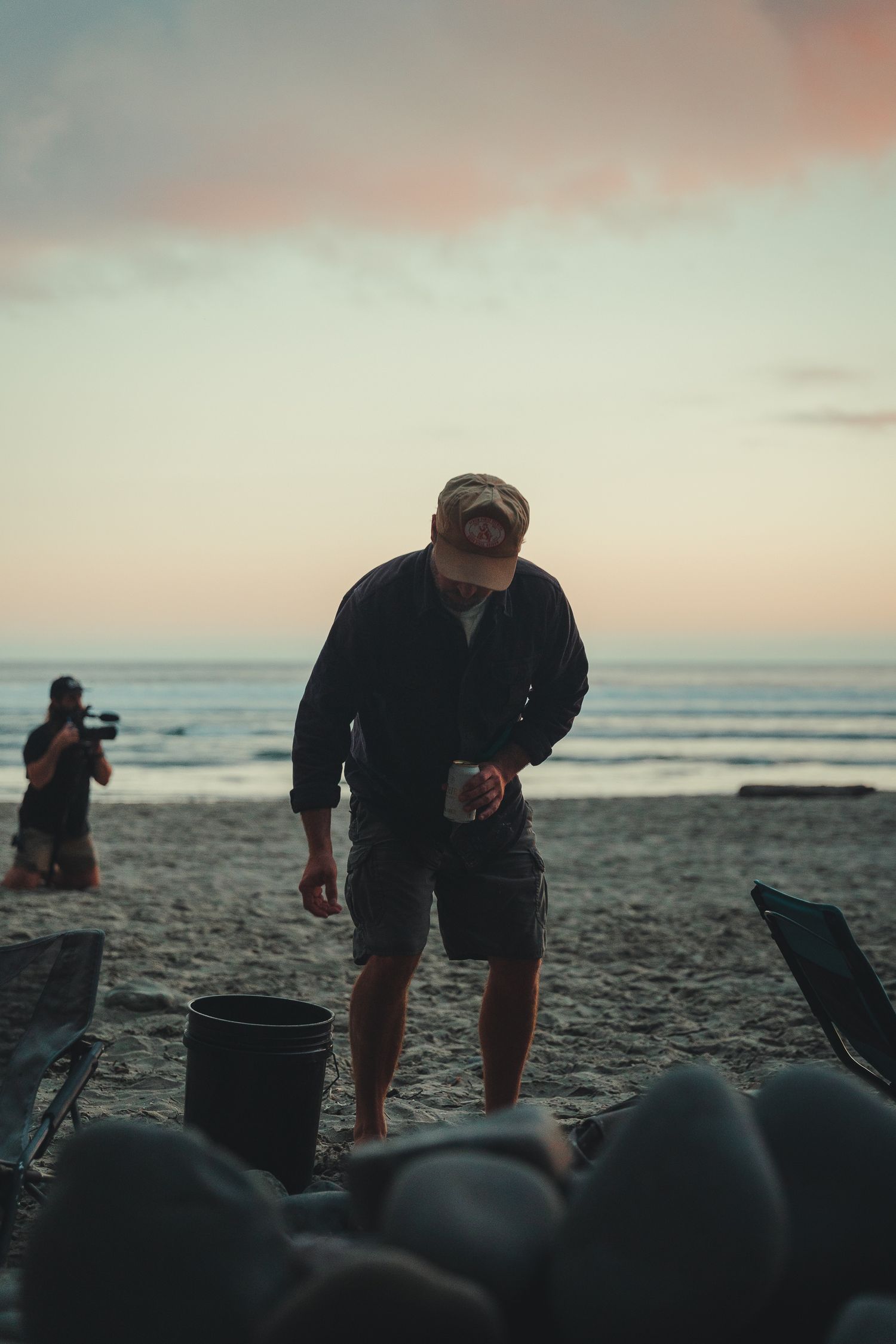 Man in cap and shorts working on a beach at sunset with a photographer and ocean waves in the background.