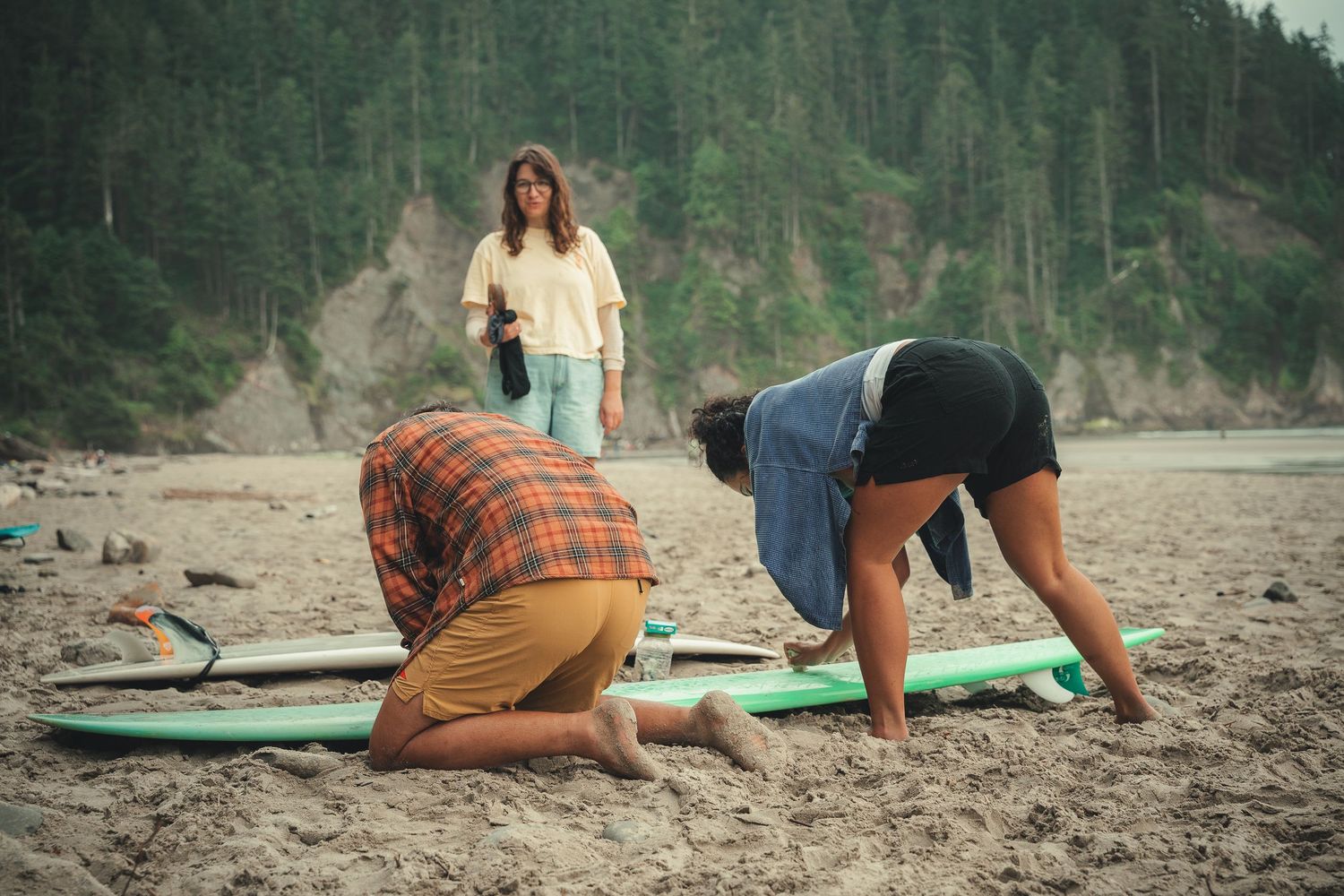 Three people on a sandy beach with a green surfboard, dense forest in the background.