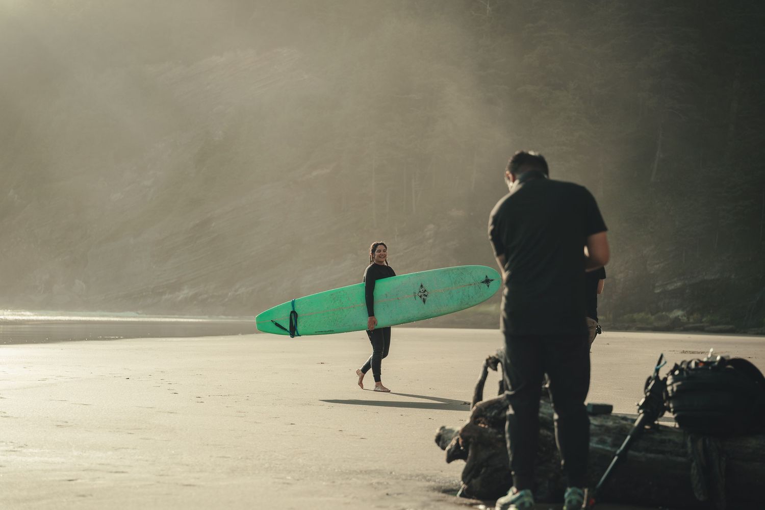 A surfer carries a green surfboard on a misty beach while a photographer with equipment captures the scene.