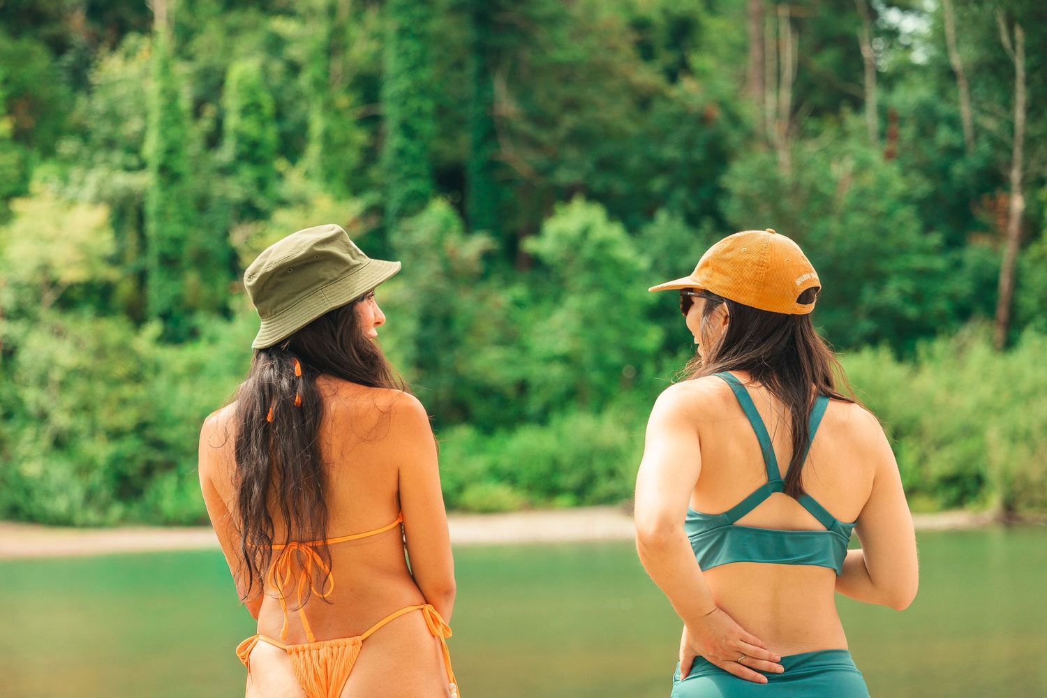 Two women in swimwear and bucket hats stand by a lush green river, viewed from behind.