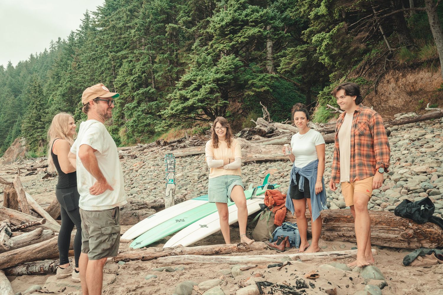 Group of friends gathered on a rocky beach with surfboards and gear, surrounded by driftwood and forest.