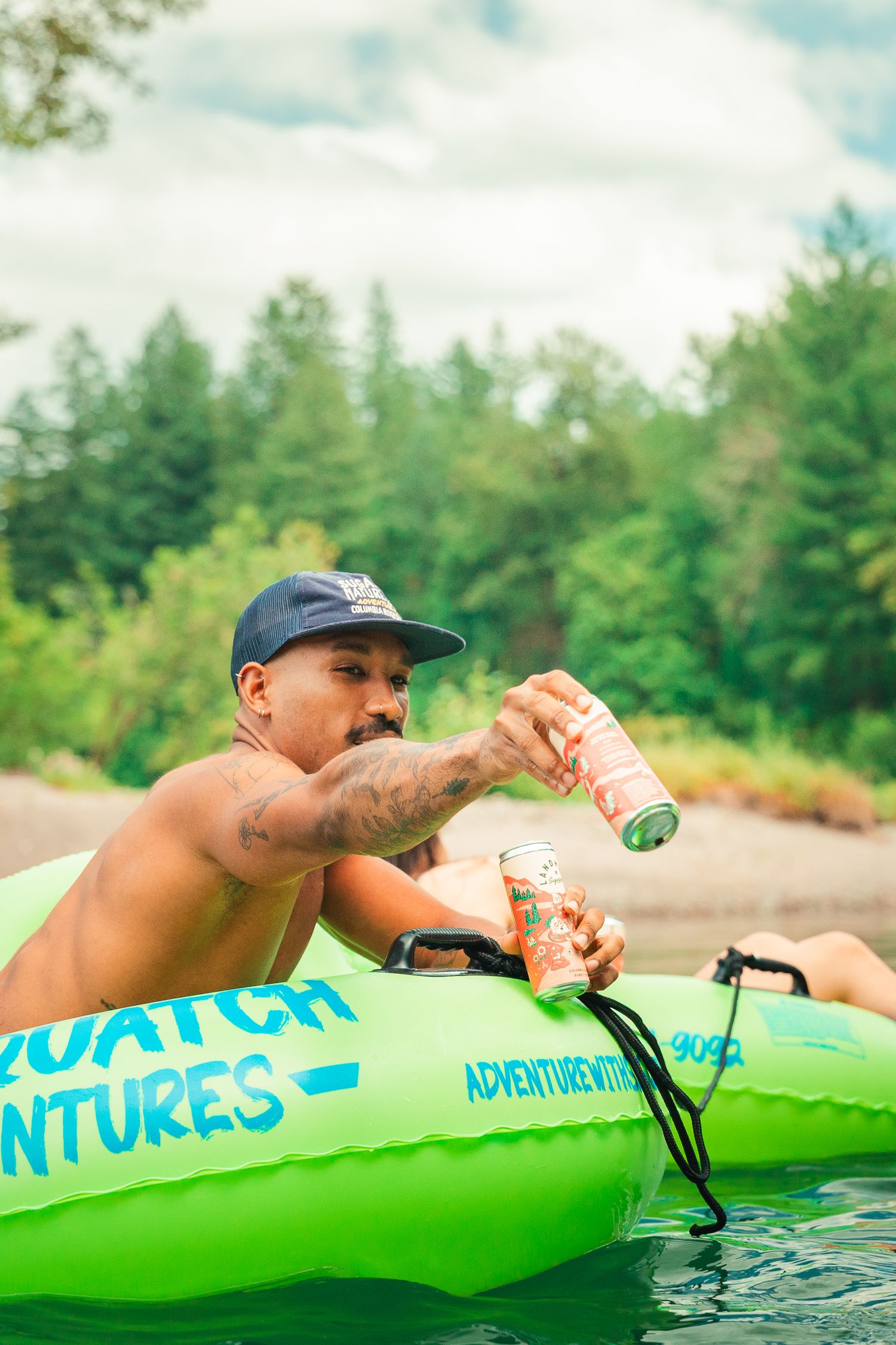 Man relaxing on green inflatable raft holding canned drinks on a lake surrounded by lush forest.